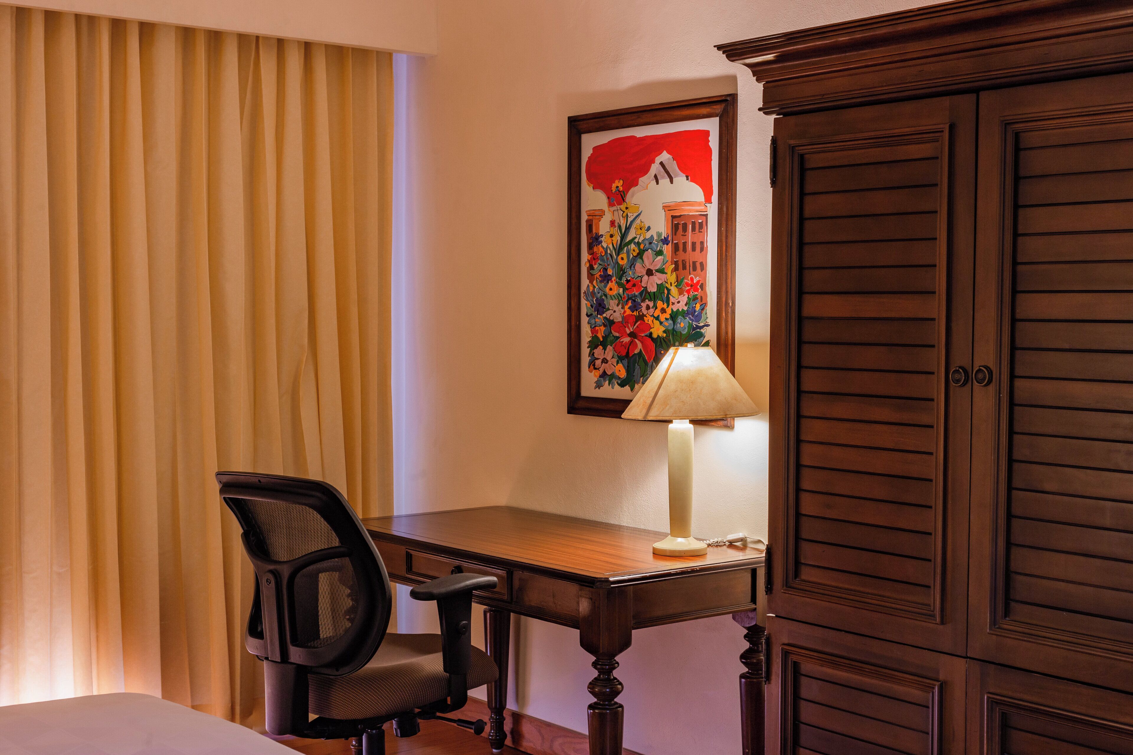 A hotel room with a wooden desk, a chair, a lamp, a bed, and a painting on the wall.