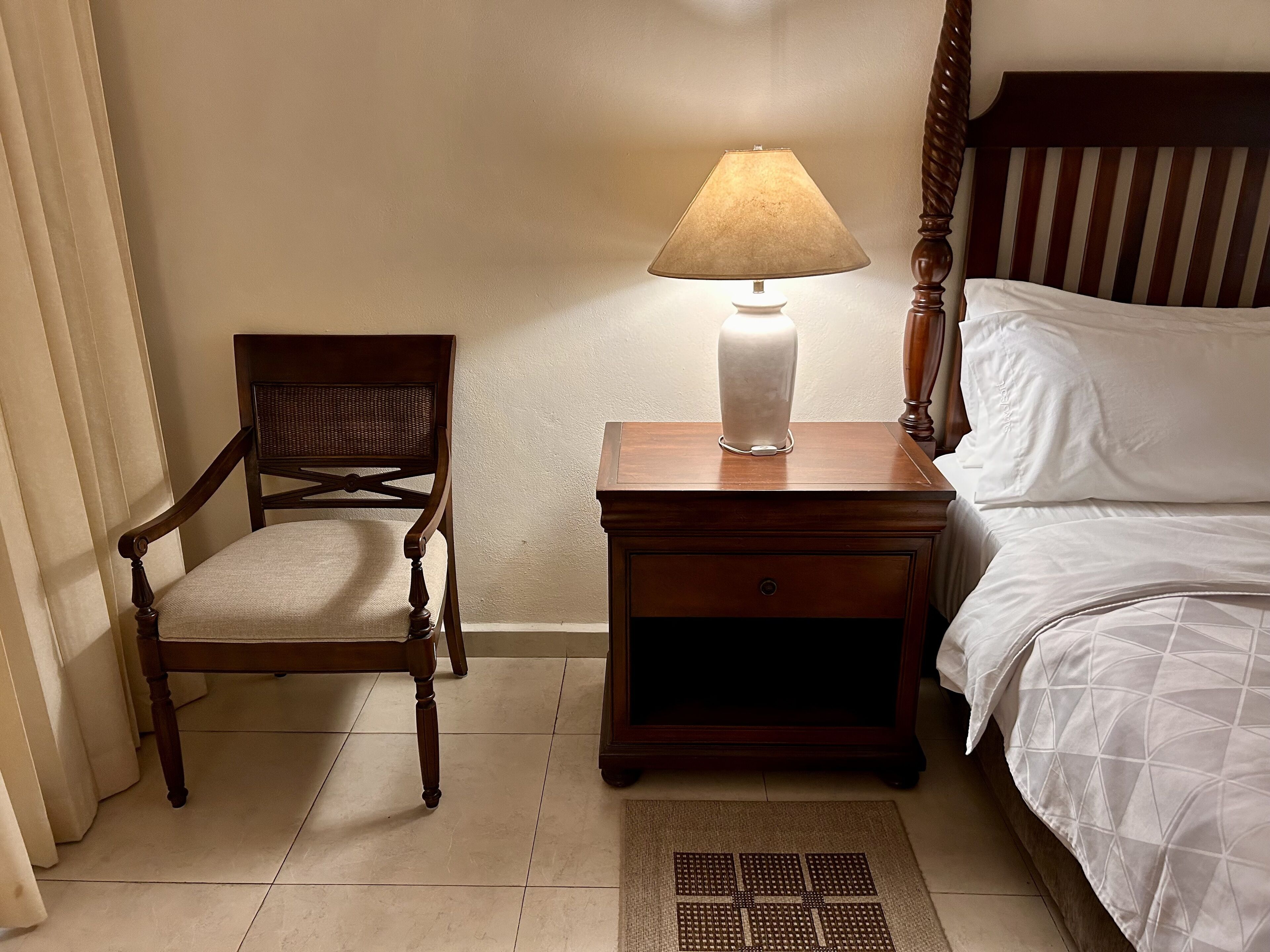 A neatly arranged bedroom with a wooden nightstand, a lamp, a chair, and a bed with white bedding.
