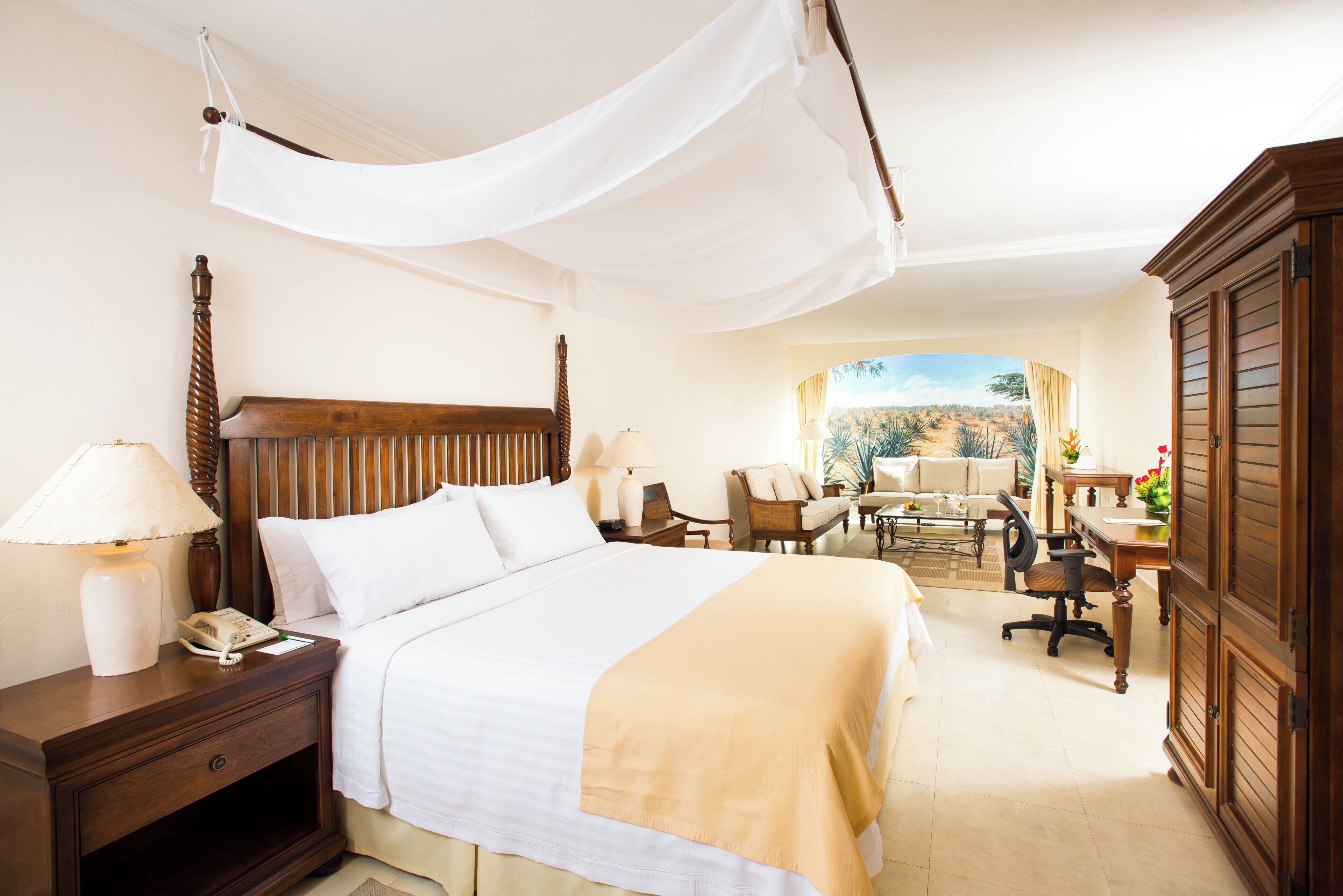A hotel room with a bed, bedside tables, a desk, a chair, a wardrobe, and a view of a desert landscape.