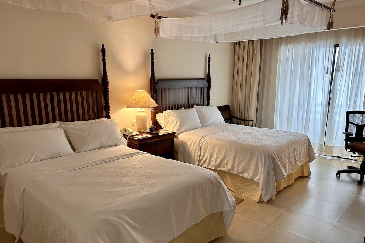 Habitación de hotel con dos camas, una mesita de noche con lámpara, una silla y una ventana con cortinas.