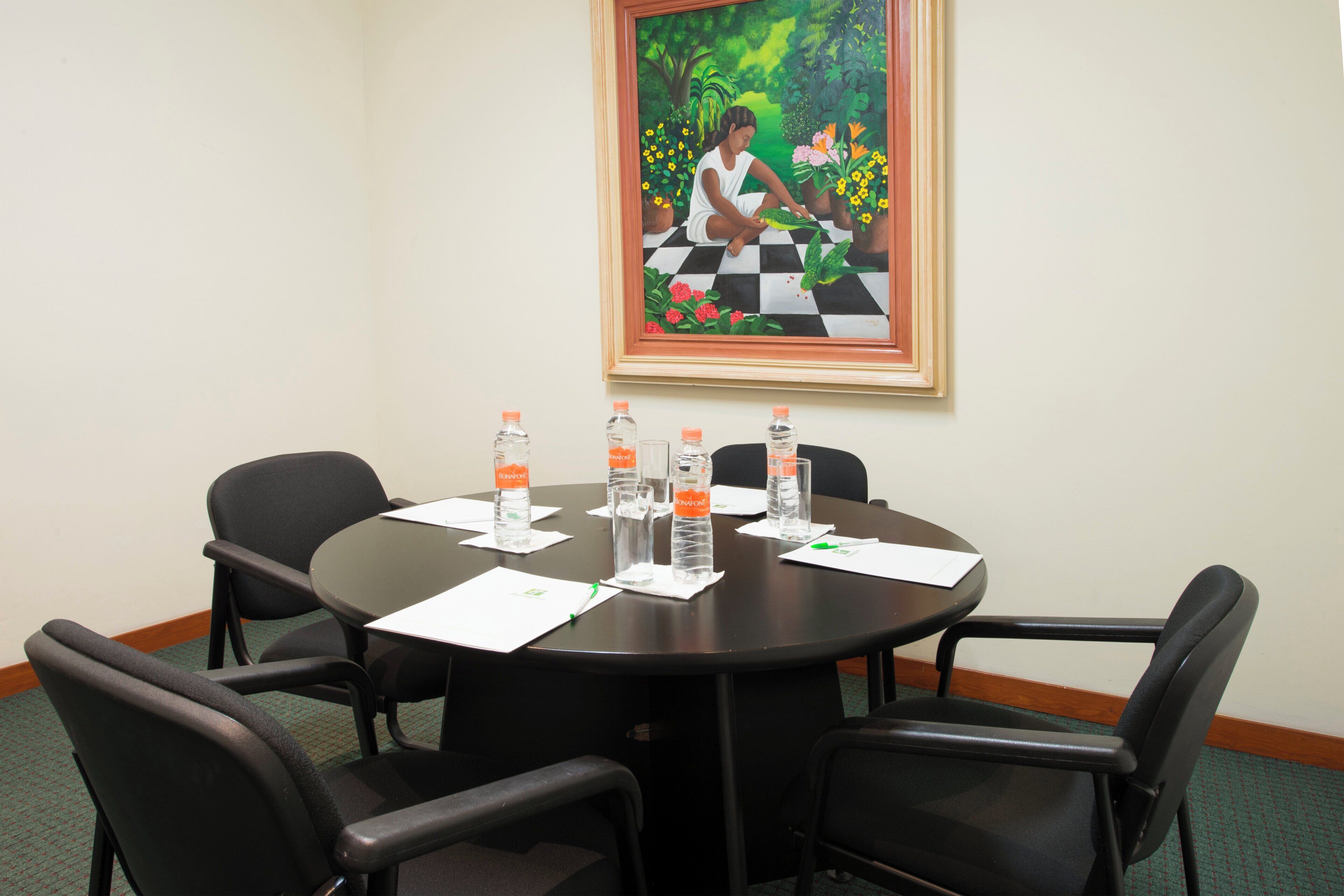 A conference room with a round table, four chairs, water bottles, and a framed painting on the wall.