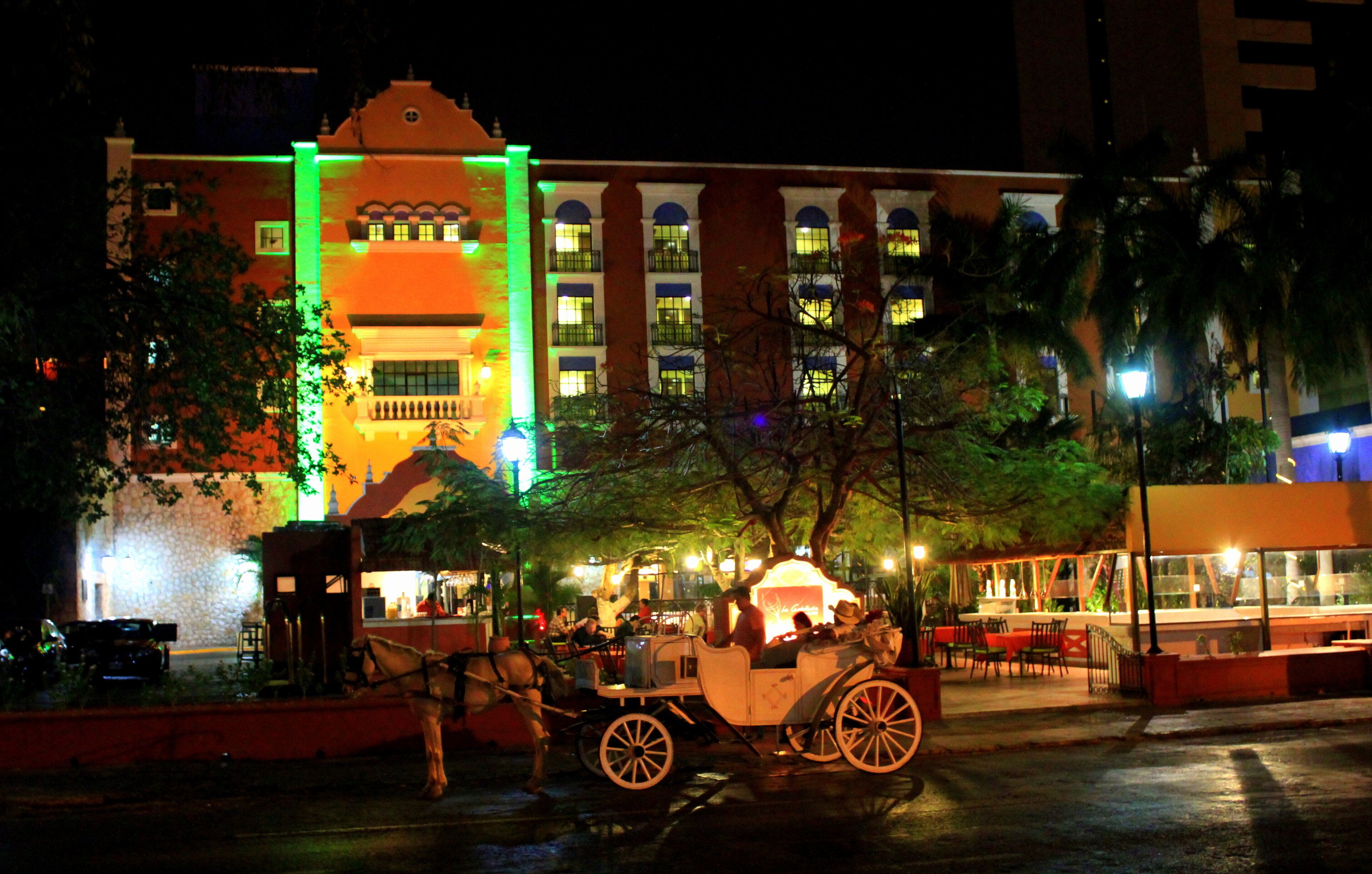 Scena notturna con un edificio illuminato da luci verdi e gialle, una carrozza trainata da cavalli e aree salotto all'aperto.