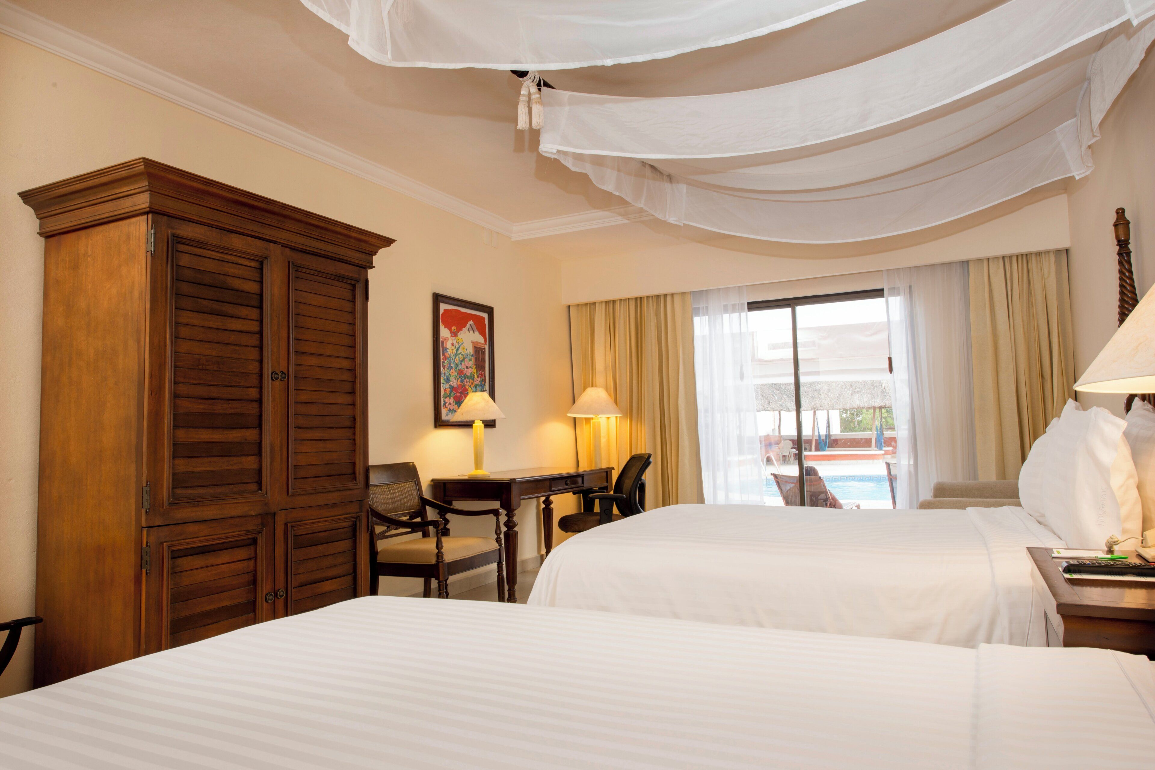 Una habitación de hotel con dos camas, un armario de madera, un escritorio con una silla, un cuadro en la pared y vista a una piscina por la ventana.