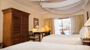 Una habitación de hotel con dos camas, un armario de madera, un escritorio con una silla, un cuadro en la pared y vista a una piscina por la ventana.