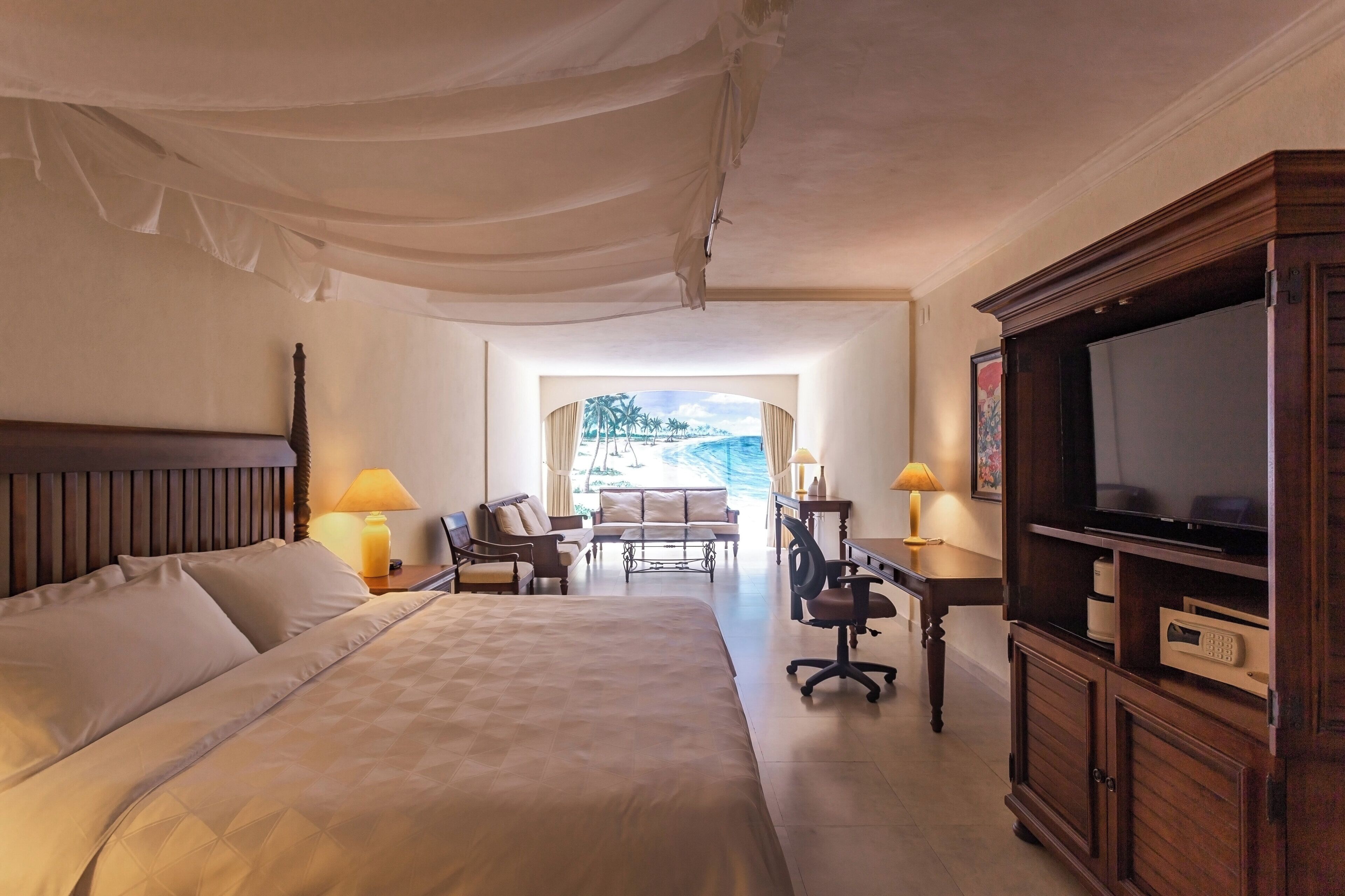 A hotel room with a large bed, a desk with a chair, a television, and a view of palm trees and a beach through the window.