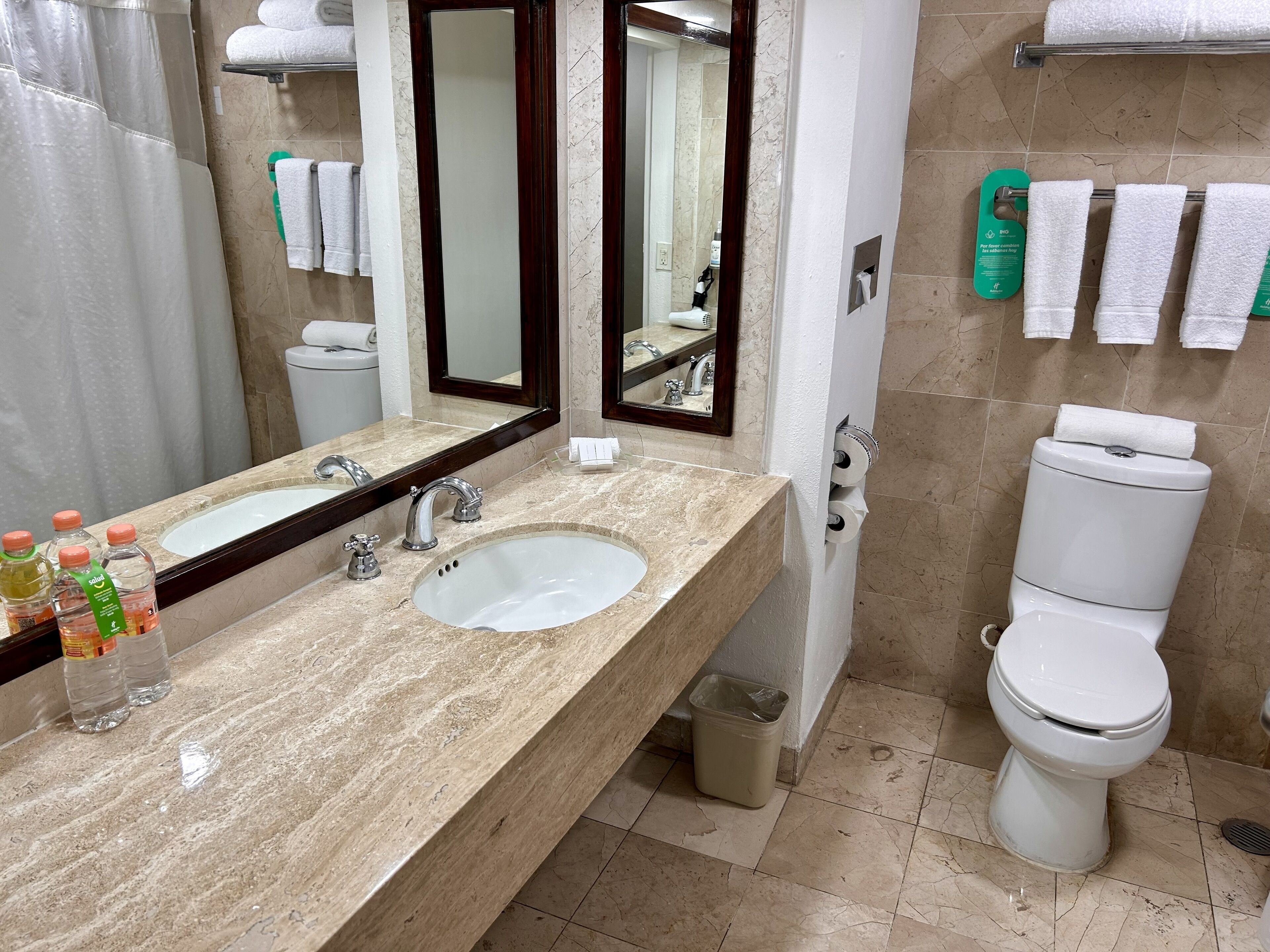 Une salle de bain avec un lavabo en marbre, deux robinets, un miroir, des toilettes et un rideau de douche.