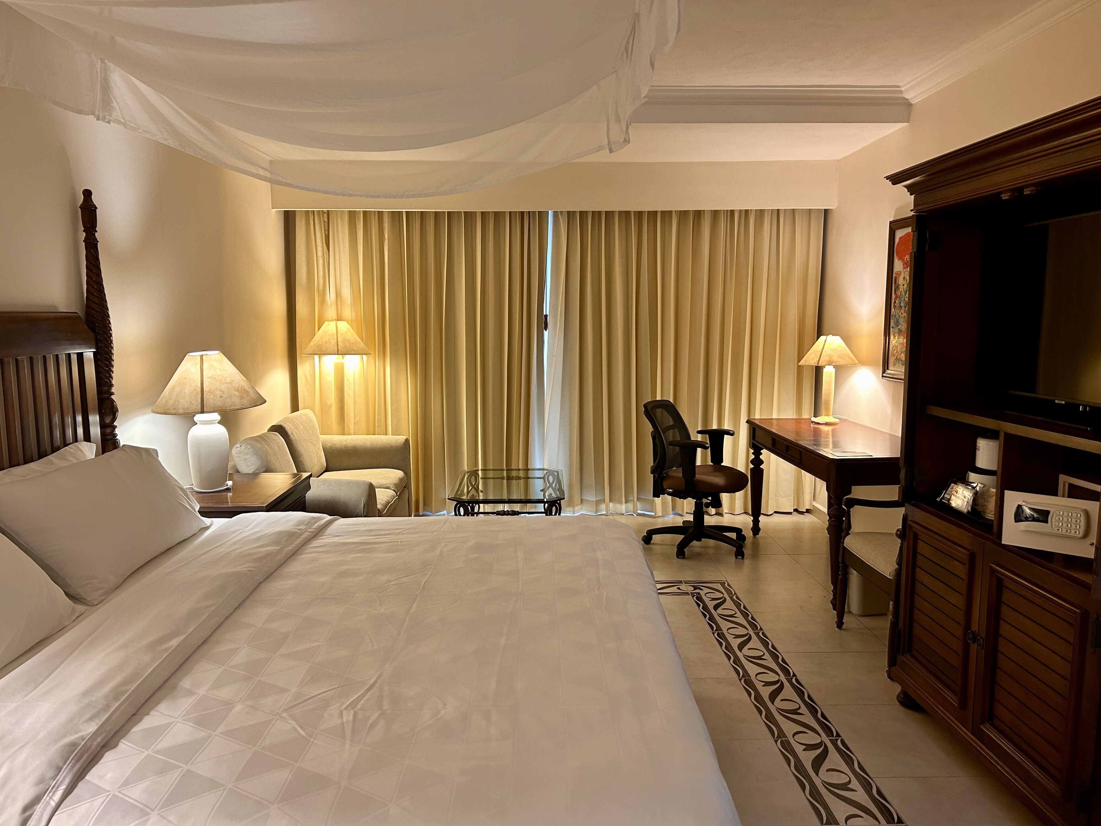 A hotel room with a large bed, two chairs, a desk, a television, and curtains.
