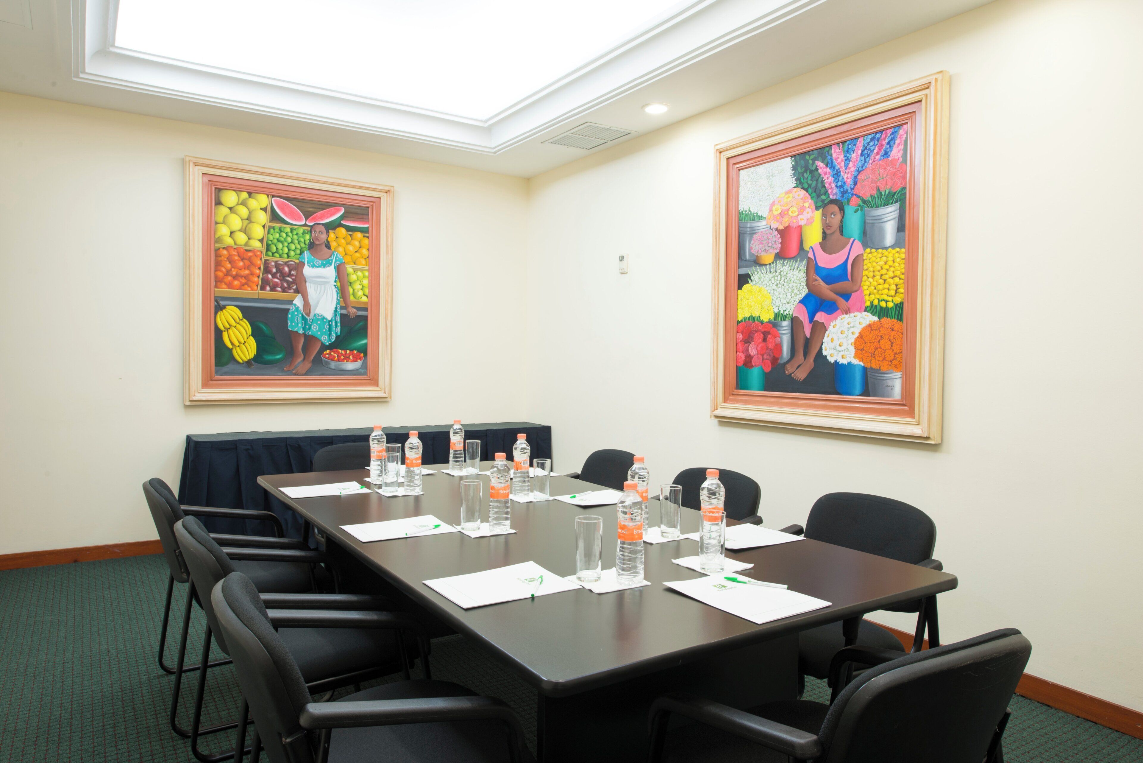 A conference room with a long table, chairs, water bottles, and papers. There are two large paintings on the walls.