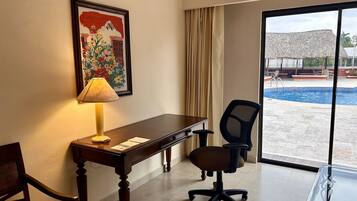 A hotel room with a desk, chair, and a large window with a view of a pool and outdoor seating.