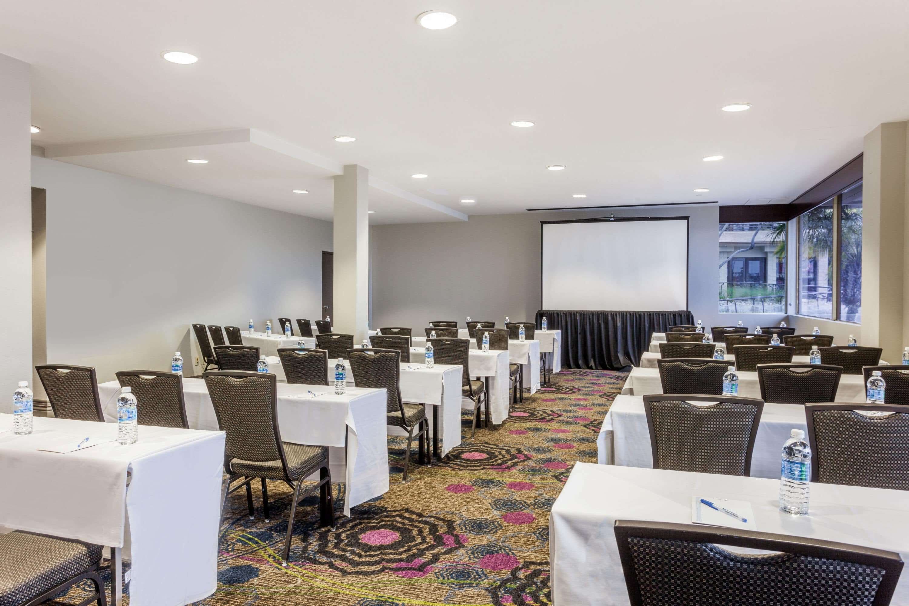 A conference room with rows of tables and chairs, water bottles on each table, and a projector screen at the front.