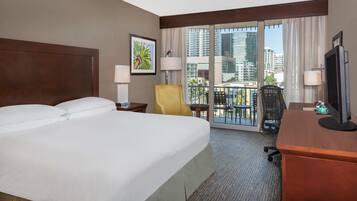 A hotel room with a large bed, a desk with a television, a chair, a balcony with a view of the city, and a window with curtains.