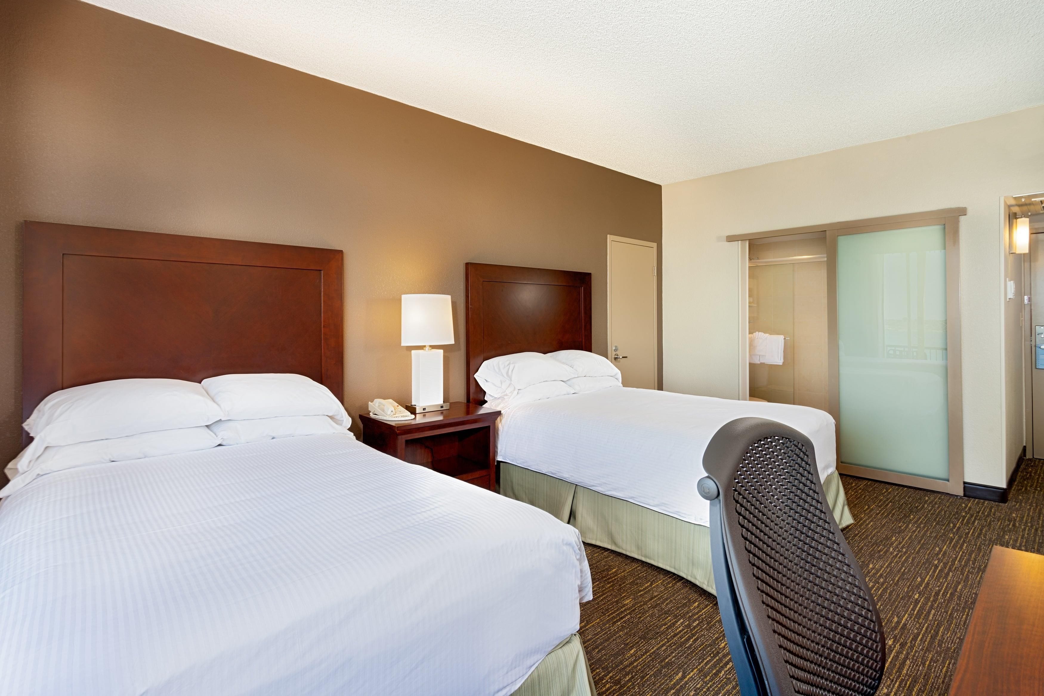 A hotel room with two beds, a nightstand with a lamp, a desk, and a bathroom with a shower and toilet.