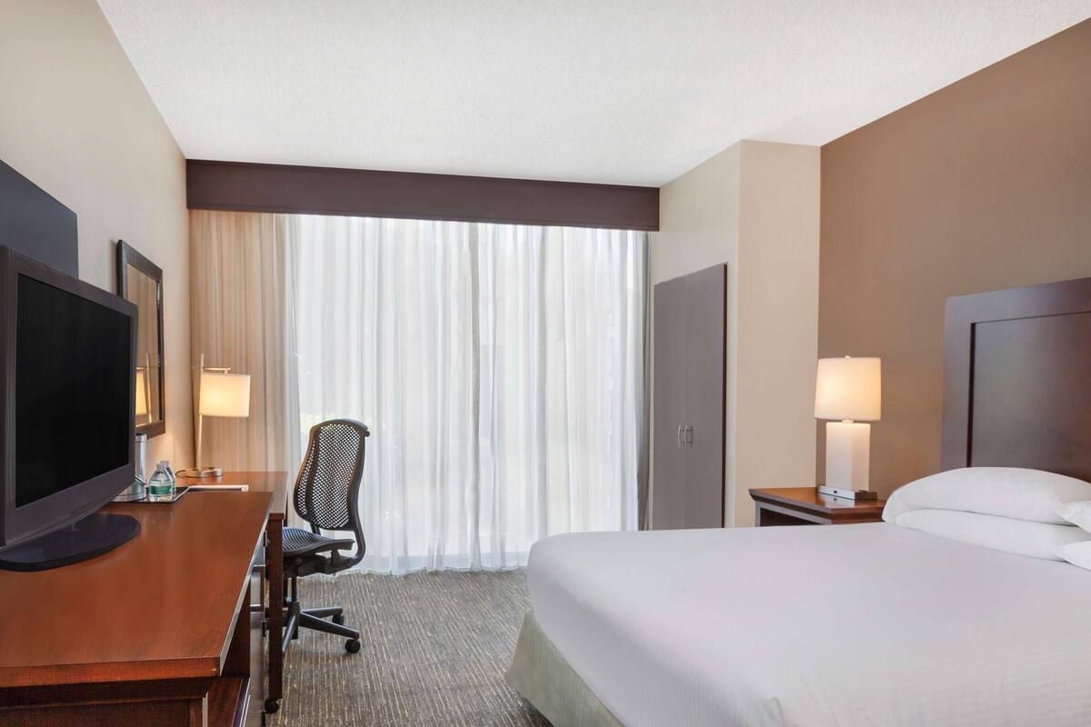 Een hotelkamer met een bed, een bureau met televisie, een stoel, een lamp en een raam met gordijnen.