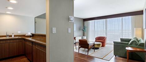 A hotel room with a kitchenette, a seating area with a sofa and armchairs, a dining table, and a large window with curtains.