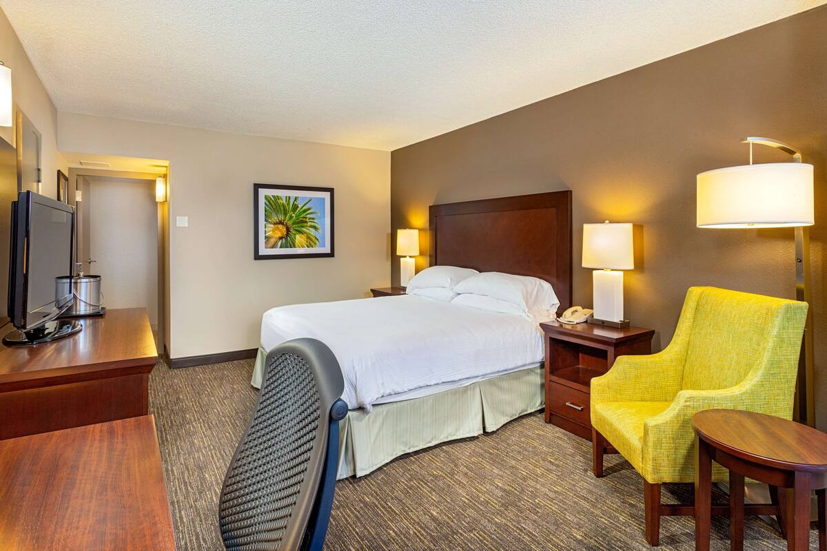Een hotelkamer met een bed, een bureau met een computer, een stoel, een lamp en een schilderij aan de muur.