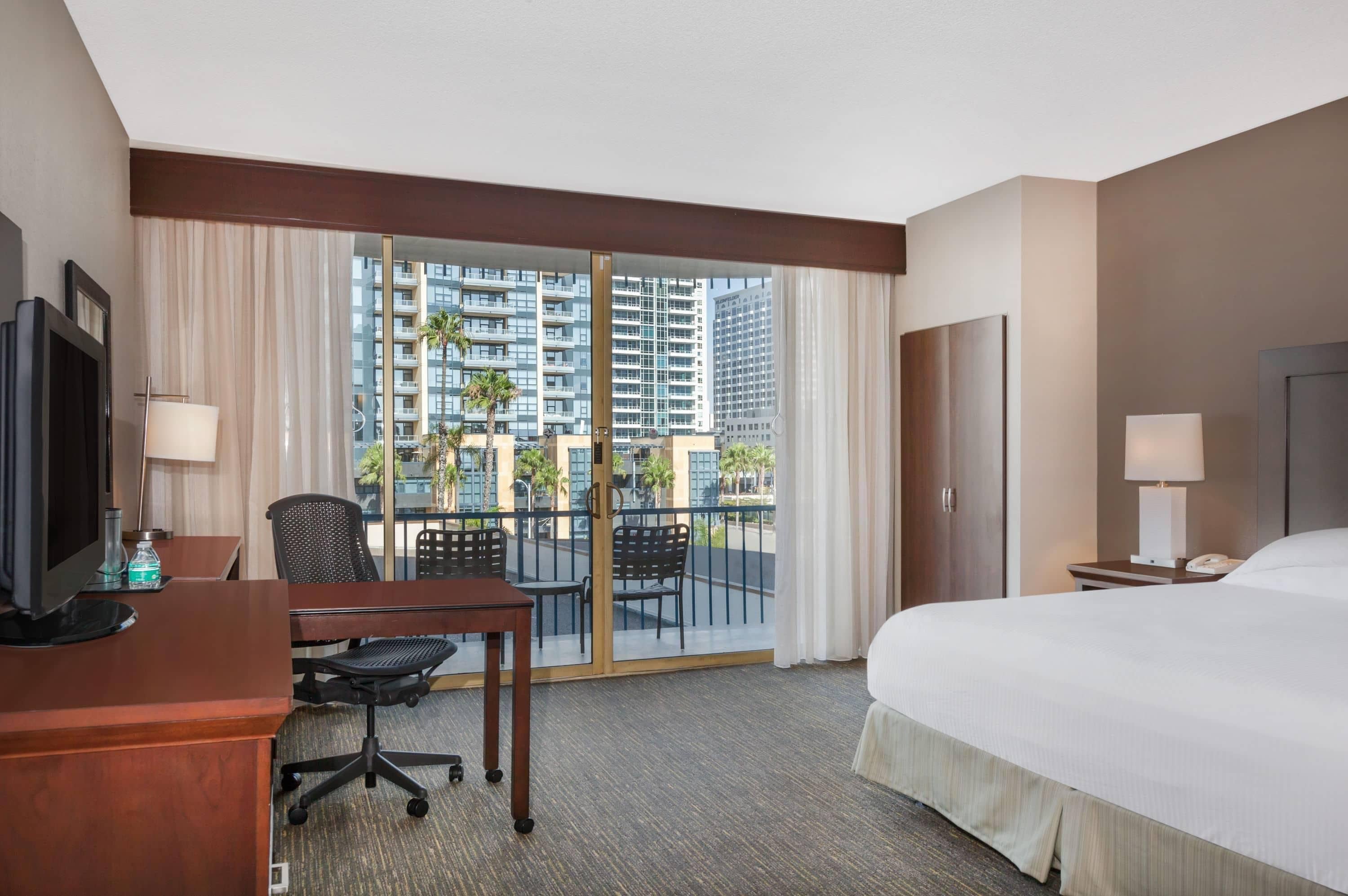 A hotel room with a bed, desk, chair, and a view of high-rise buildings.
