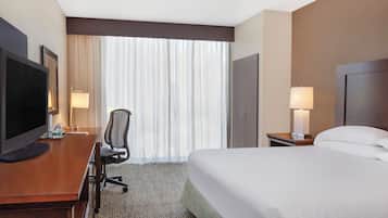 A hotel room with a bed, a desk with a television, a chair, a lamp, and a window with curtains.