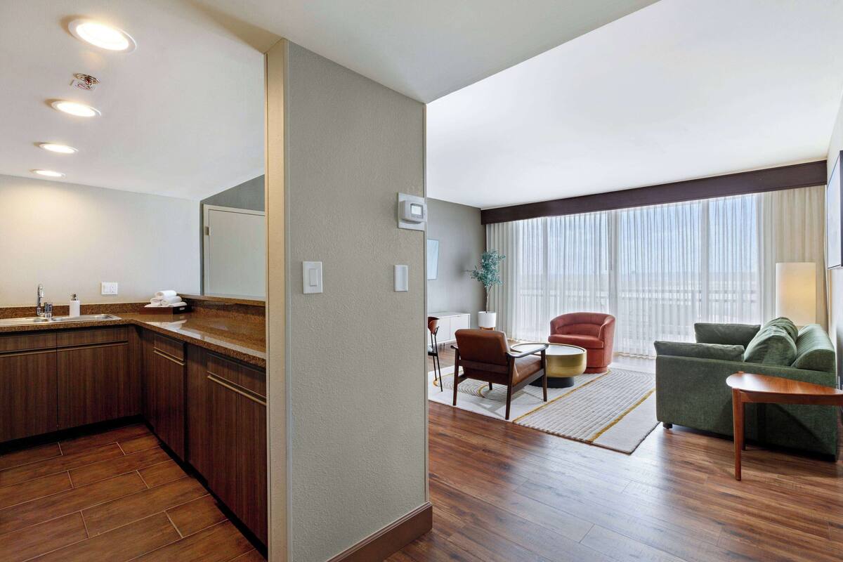 Hotelkamer met kitchenette, zithoek met bank en fauteuil, eettafel en een groot raam met gordijnen.