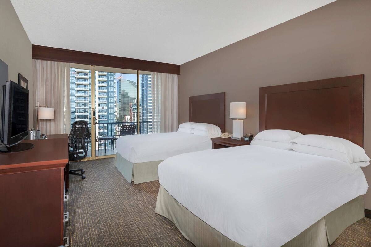 Een hotelkamer met twee bedden, een bureau, een televisie en uitzicht op een stadsbeeld door grote ramen.