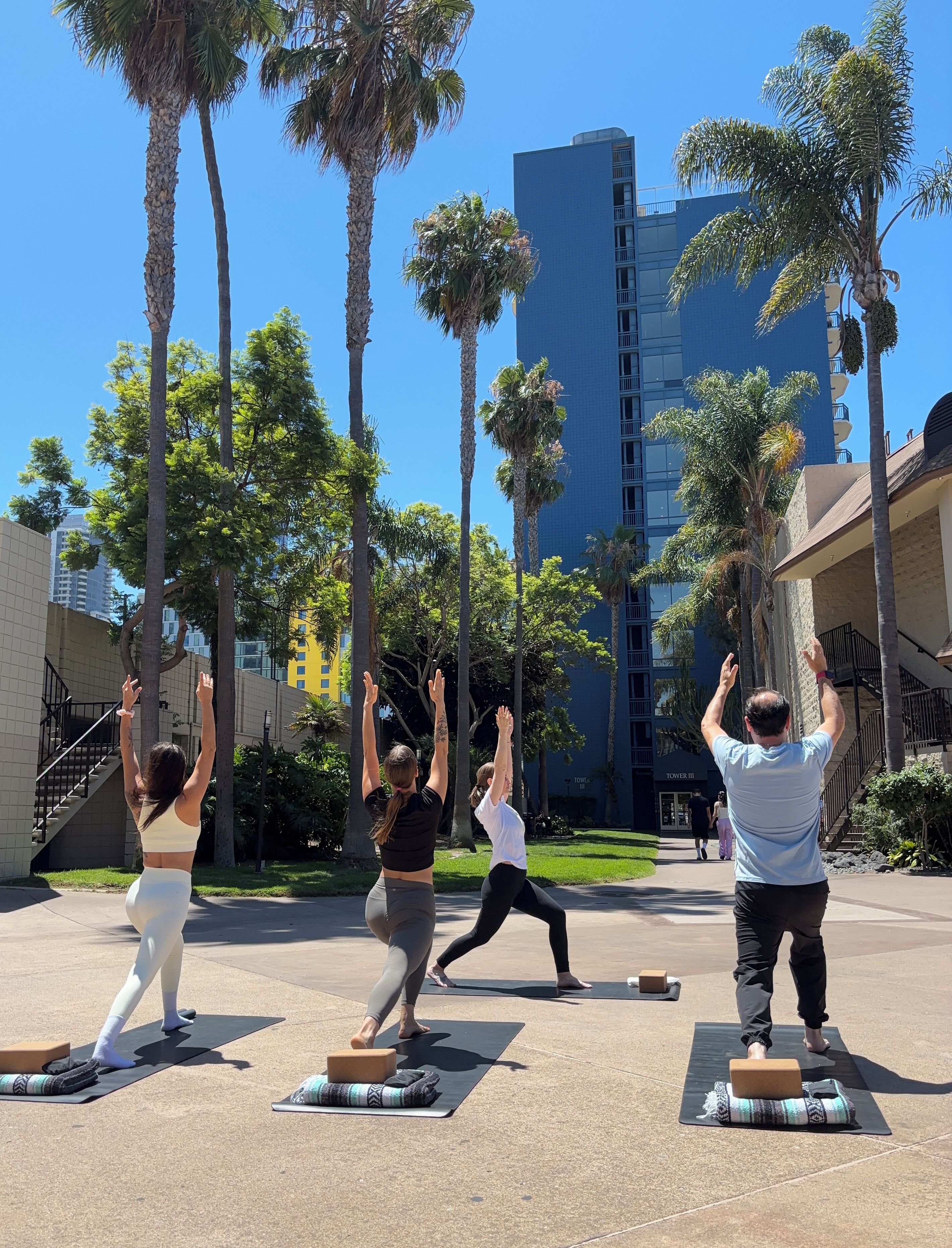 Un groupe de personnes pratiquant une posture de yoga en plein air, avec des palmiers et un bâtiment en arrière-plan.