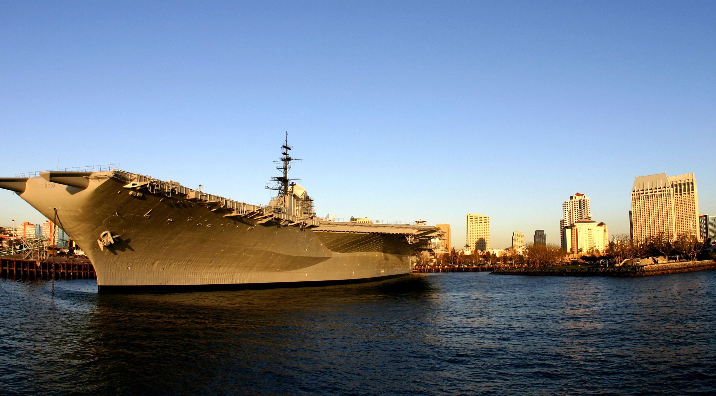 An aircraft carrier docked at a port with a city skyline in the background.