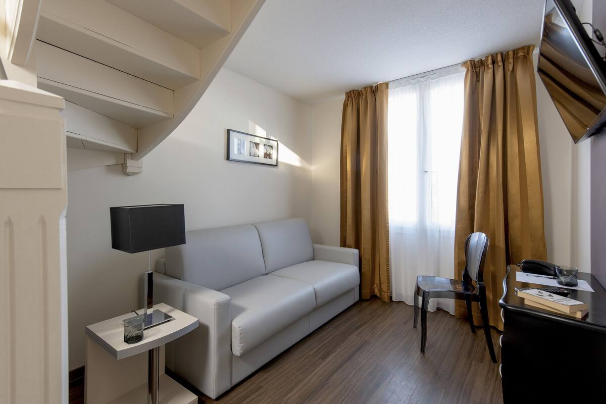 A modern hotel room with a sofa, a small table with a lamp, a chair, a desk with a phone and papers, and a window with curtains.