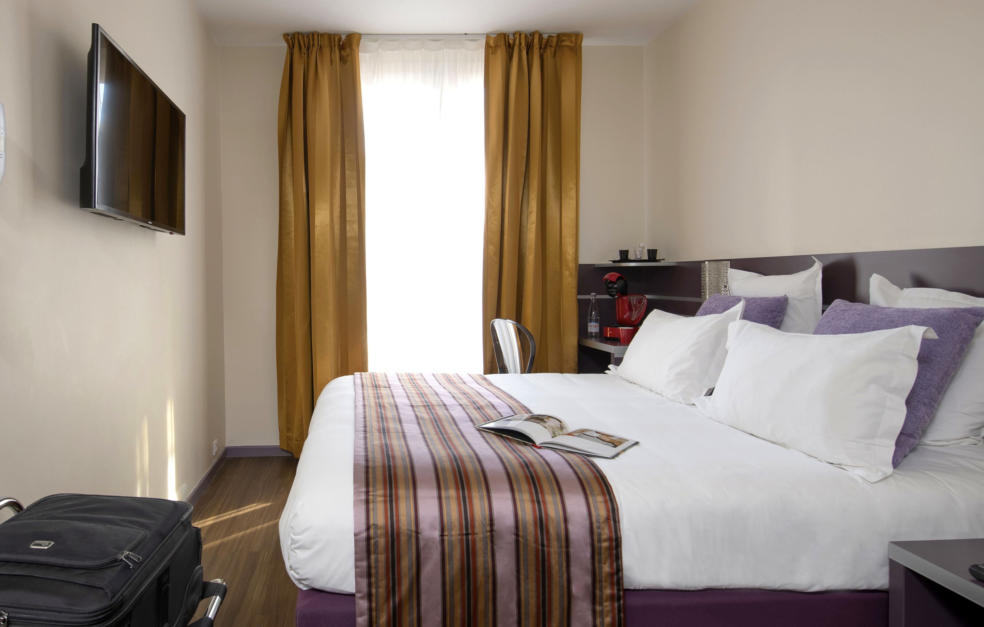 Una habitación de hotel con cama, televisión montada en la pared, maleta y ventana con cortinas.
