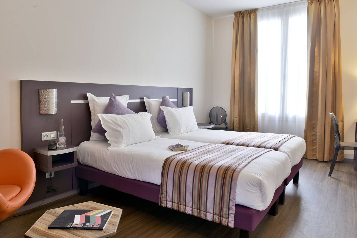 A hotel room with a large bed, a nightstand, a chair, and a bedside table.
