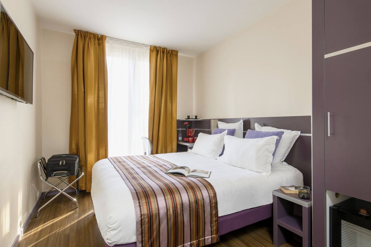 A hotel room with a bed, a bedside table, a chair, a suitcase, and a TV.