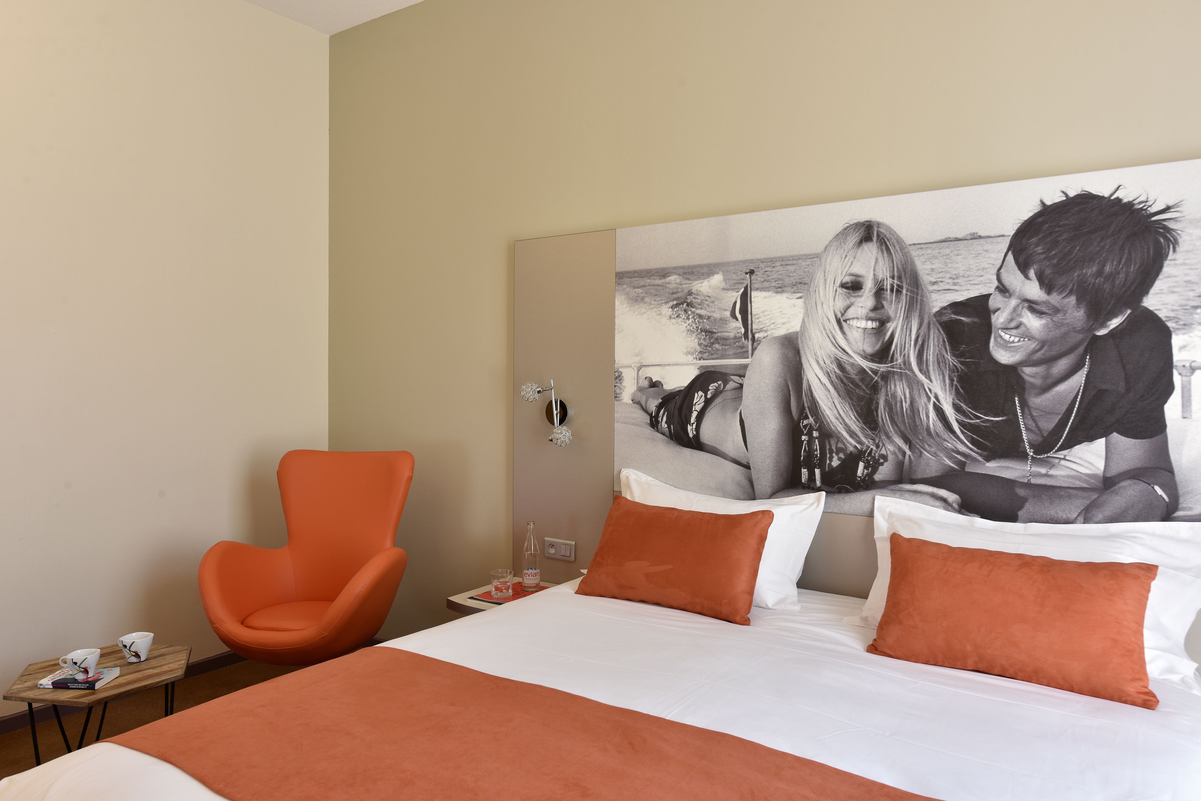 A hotel room with a large bed, orange pillows, a single orange chair, a small table with a book and a glass, and a wall-mounted picture of a couple.