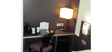 Una habitación de hotel con escritorio, una silla, lámpara, cesto de basura y cama.