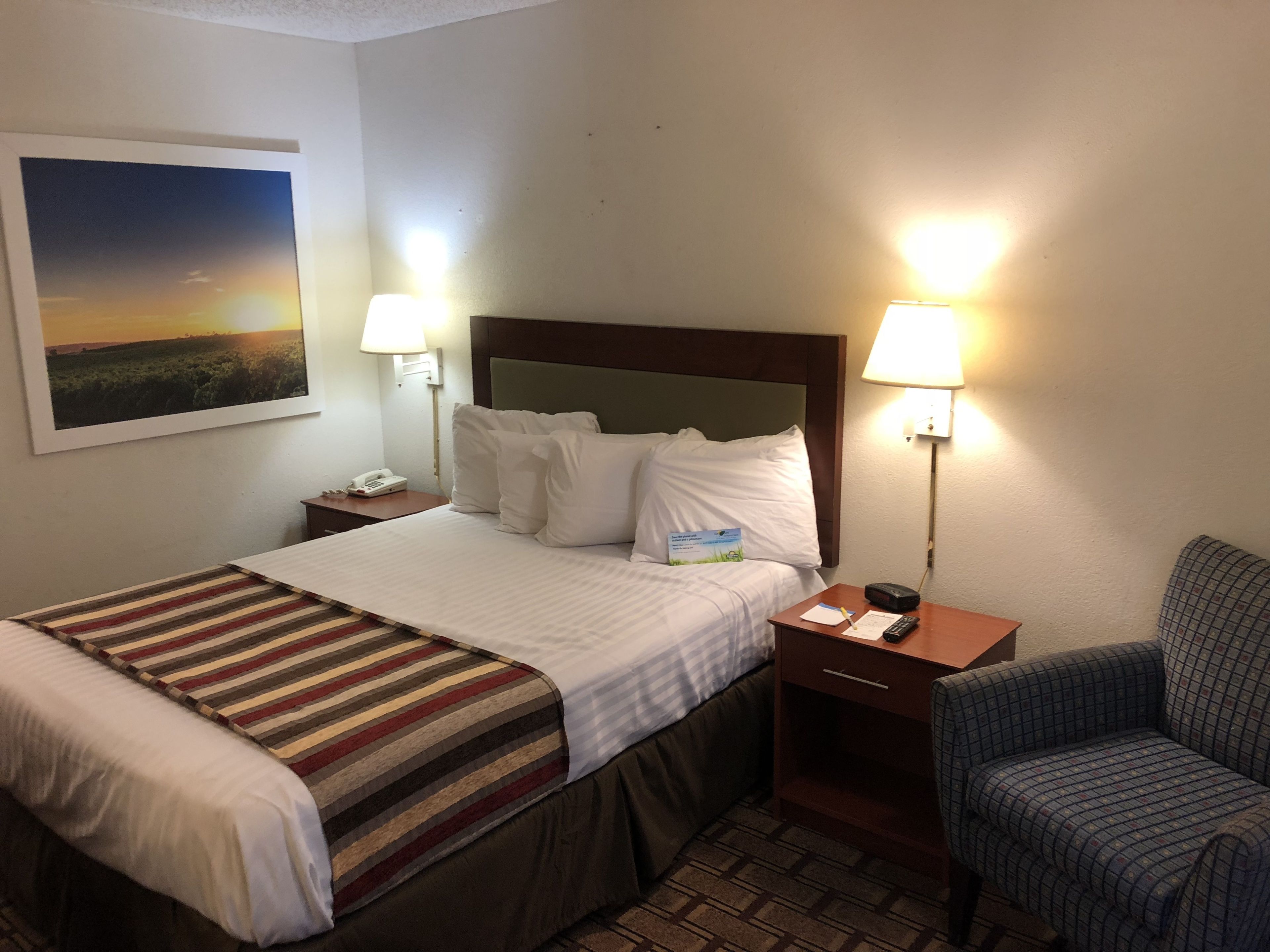 A hotel room with a bed, two bedside lamps, a nightstand with a phone, a chair, and a painting on the wall.