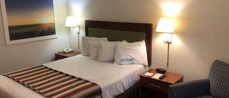 A hotel room with a bed, two bedside lamps, a nightstand with a phone, a chair, and a painting on the wall.