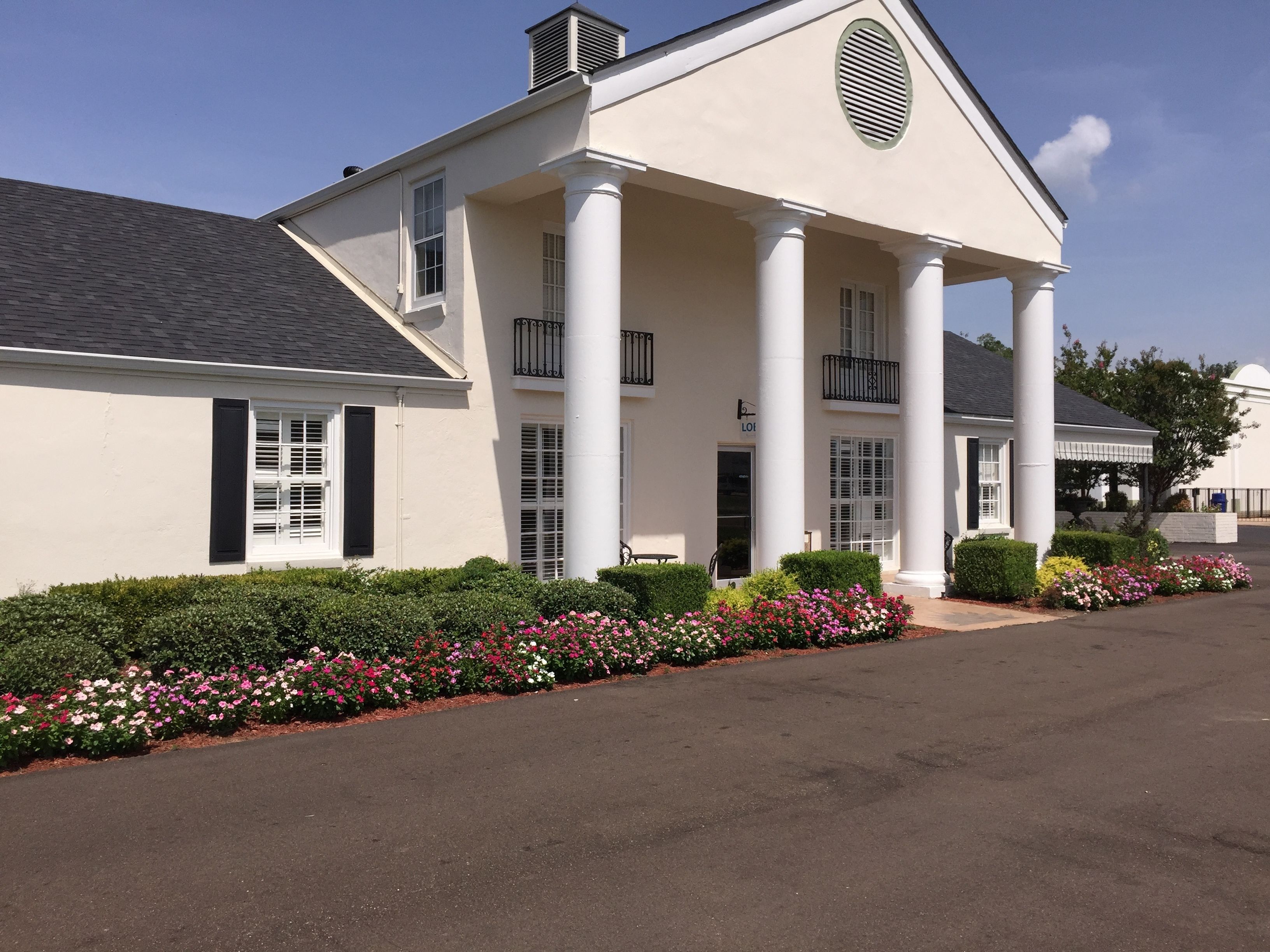 A large, white building with columns and a balcony, surrounded by a well-maintained garden with various flowers.