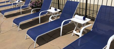 Una fila de sillones azules con estructura blanca y una mesita con una toalla, dispuesta sobre un patio de concreto con una cerca metálica negra al fondo.