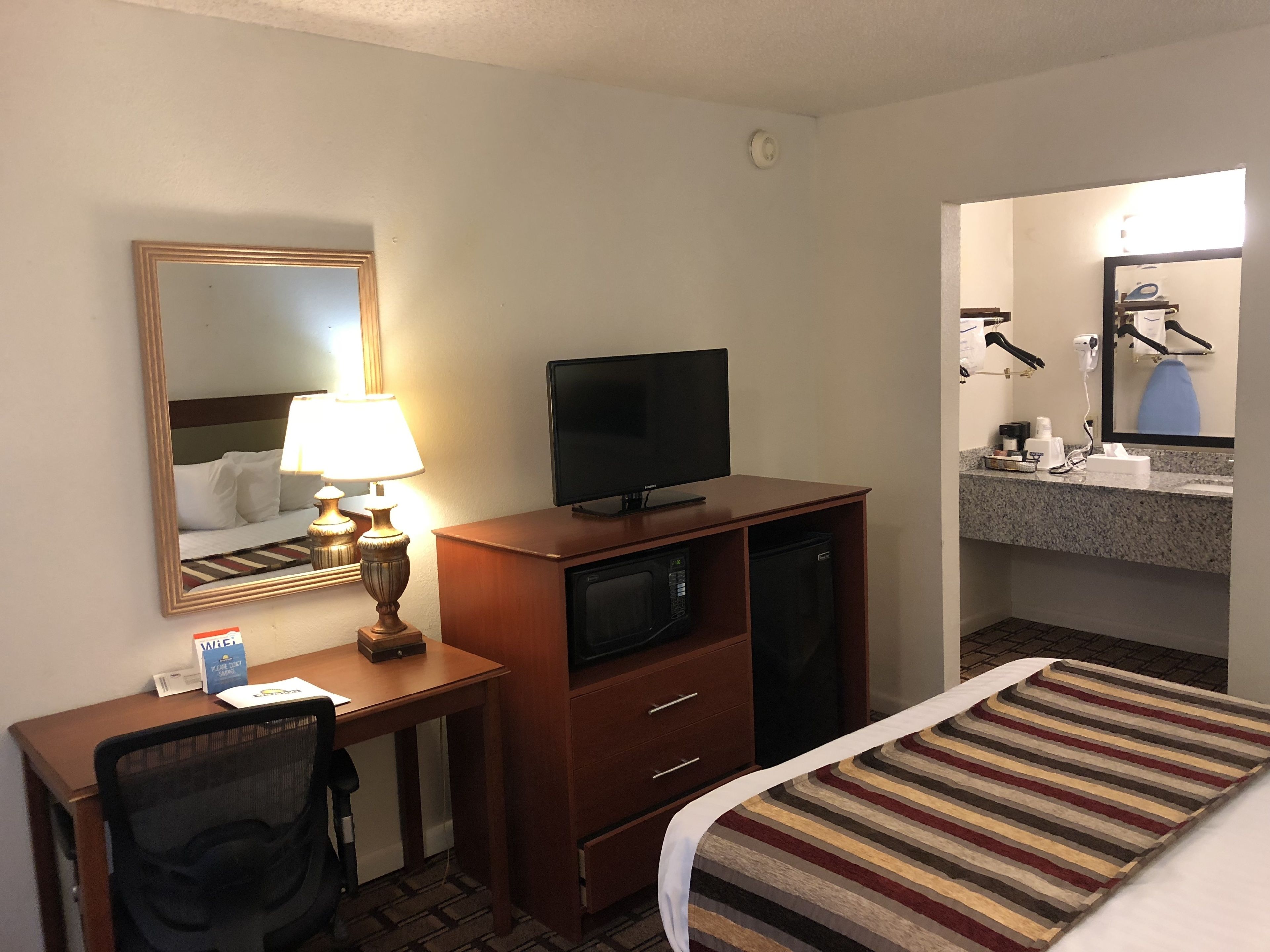 Habitación de hotel con cama, escritorio, televisor, lámpara y baño con espejo, lavabo y toallero.
