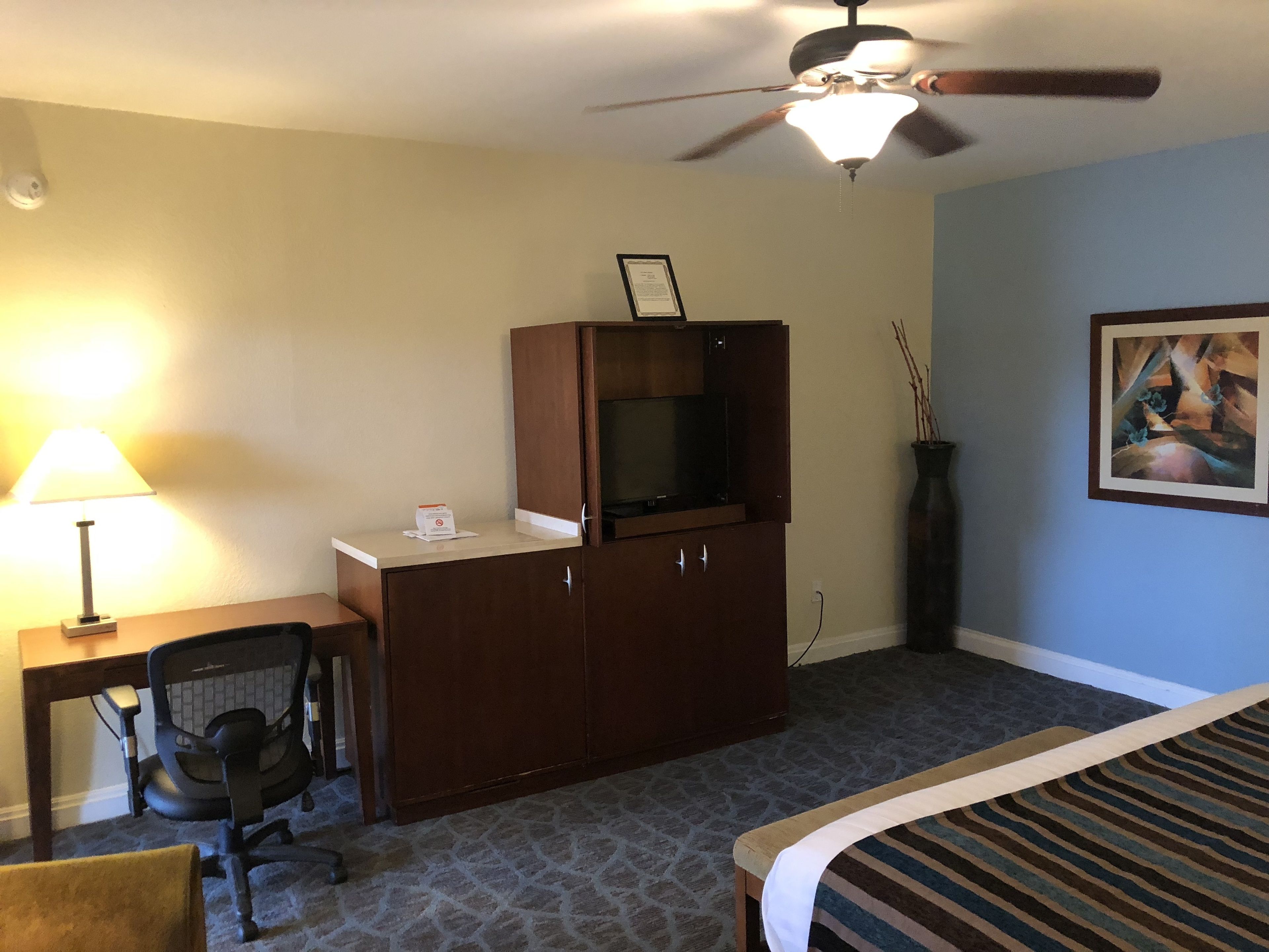A hotel room with a bed, a desk with a chair, a television on a wooden cabinet, a ceiling fan, and a painting on the wall.