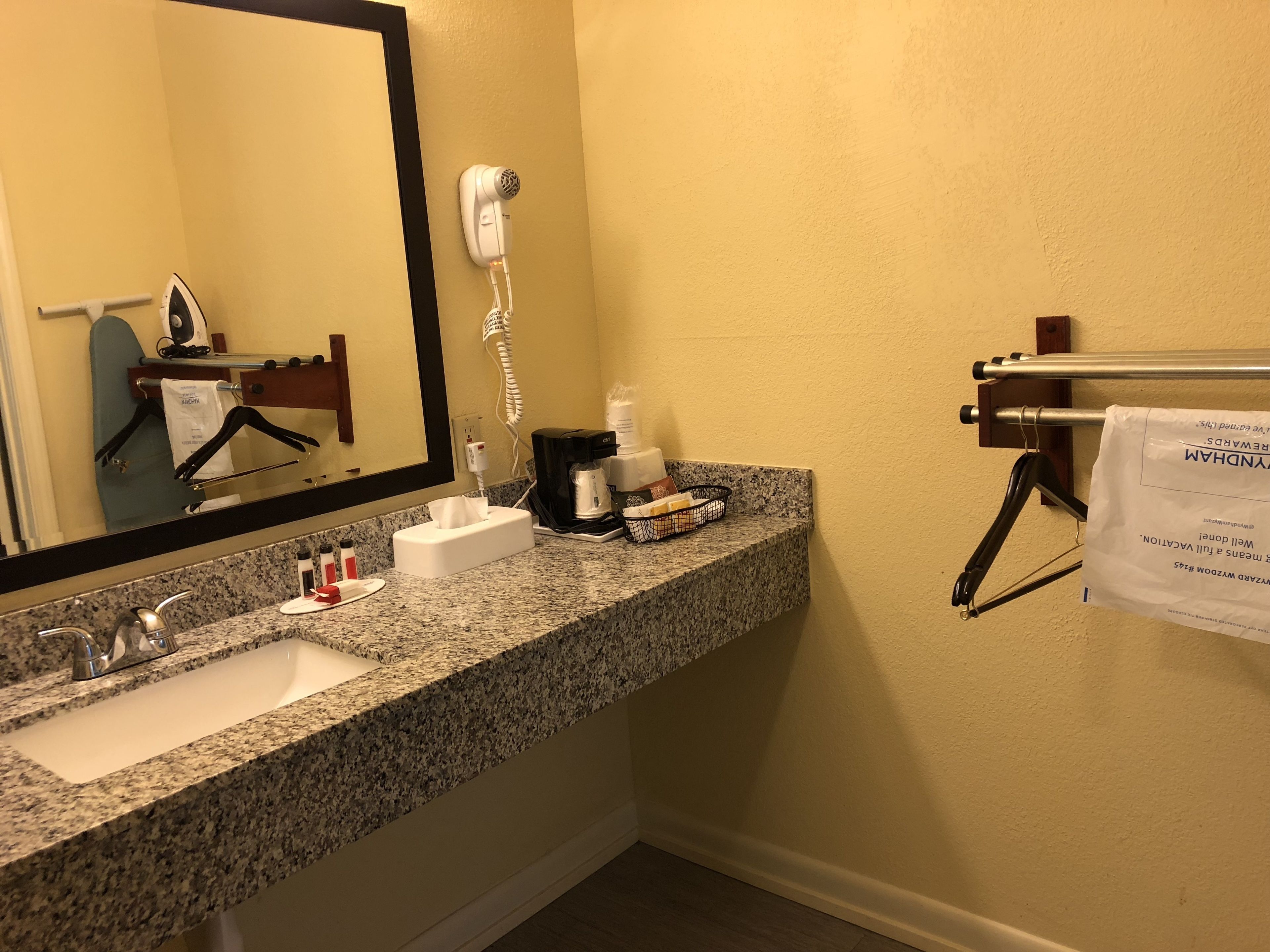 Un baño de hotel con lavamanos de encimera de granito, un secador de pelo, un perchero y un espejo.