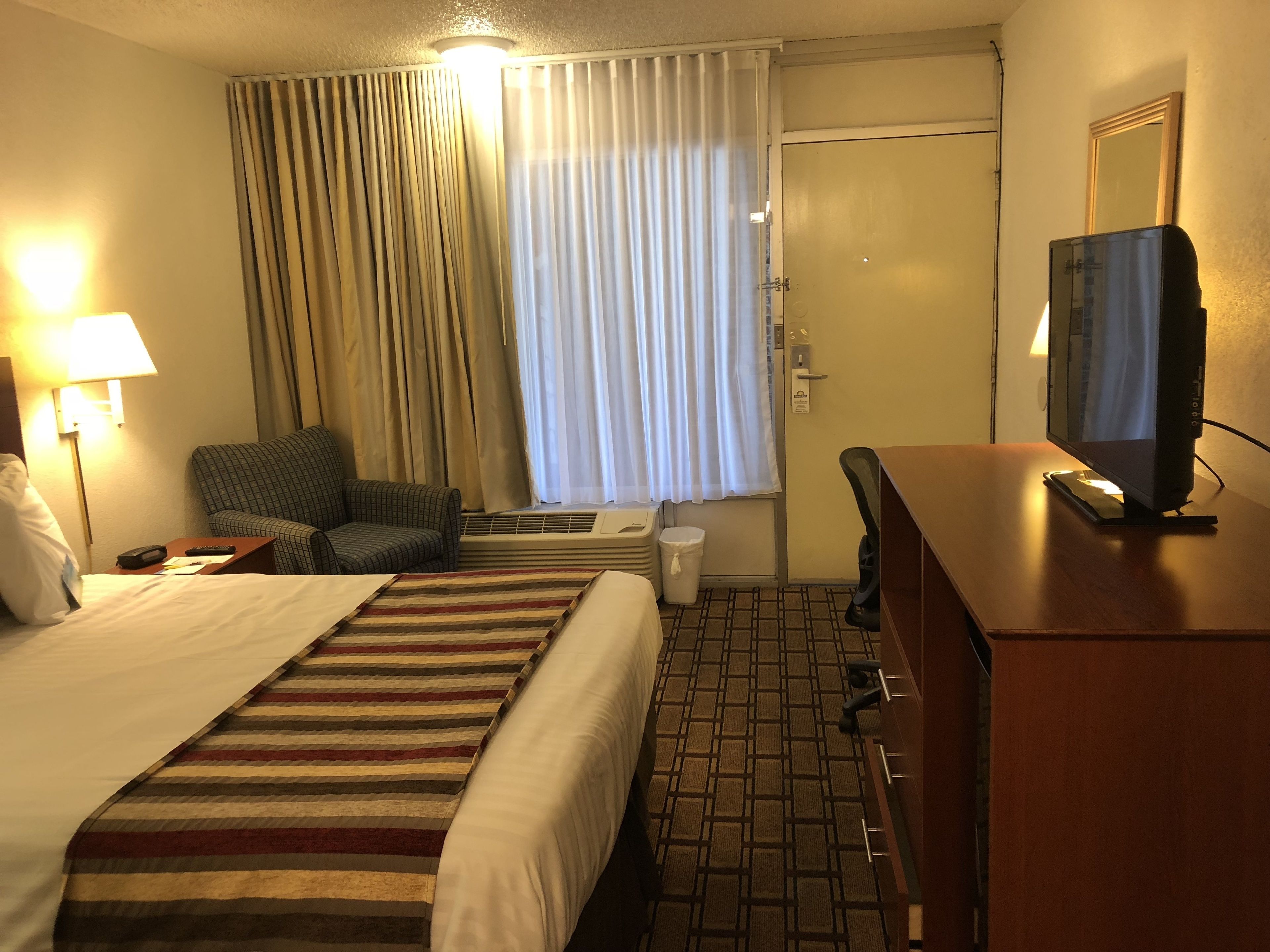 A hotel room with a bed, a chair, a desk with a television, a lamp, and a window with curtains.