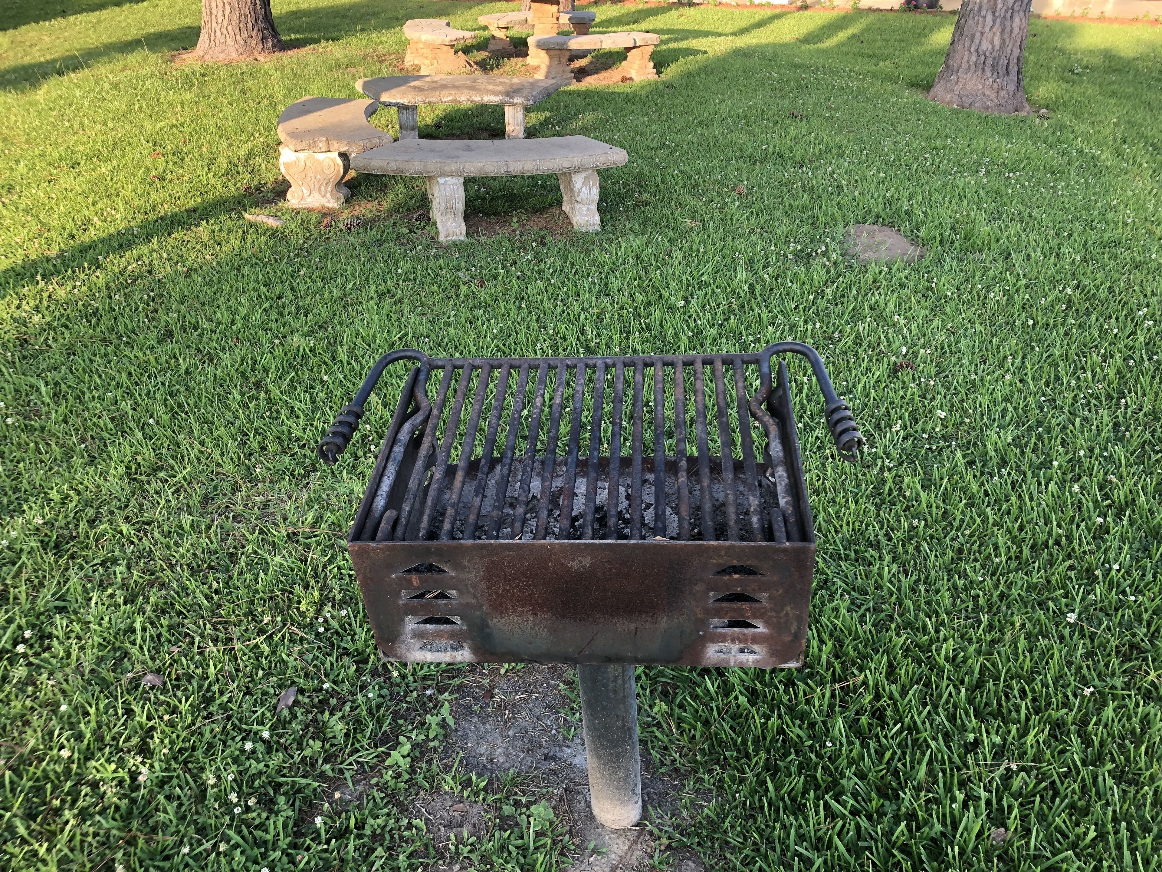 A metal barbecue grill with a flat grate and a handle on each side, placed on a grassy lawn with a stone bench and tree in the background.