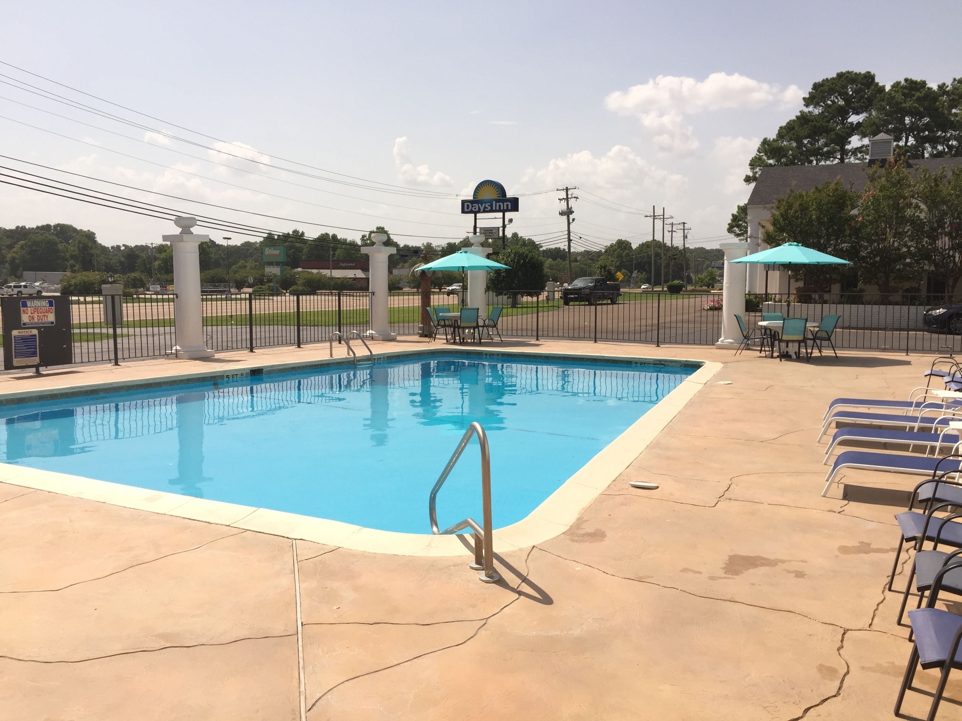 Una piscina con quiosco de salvavidas, sillas de descanso y un pabellón con sombrilla azul.