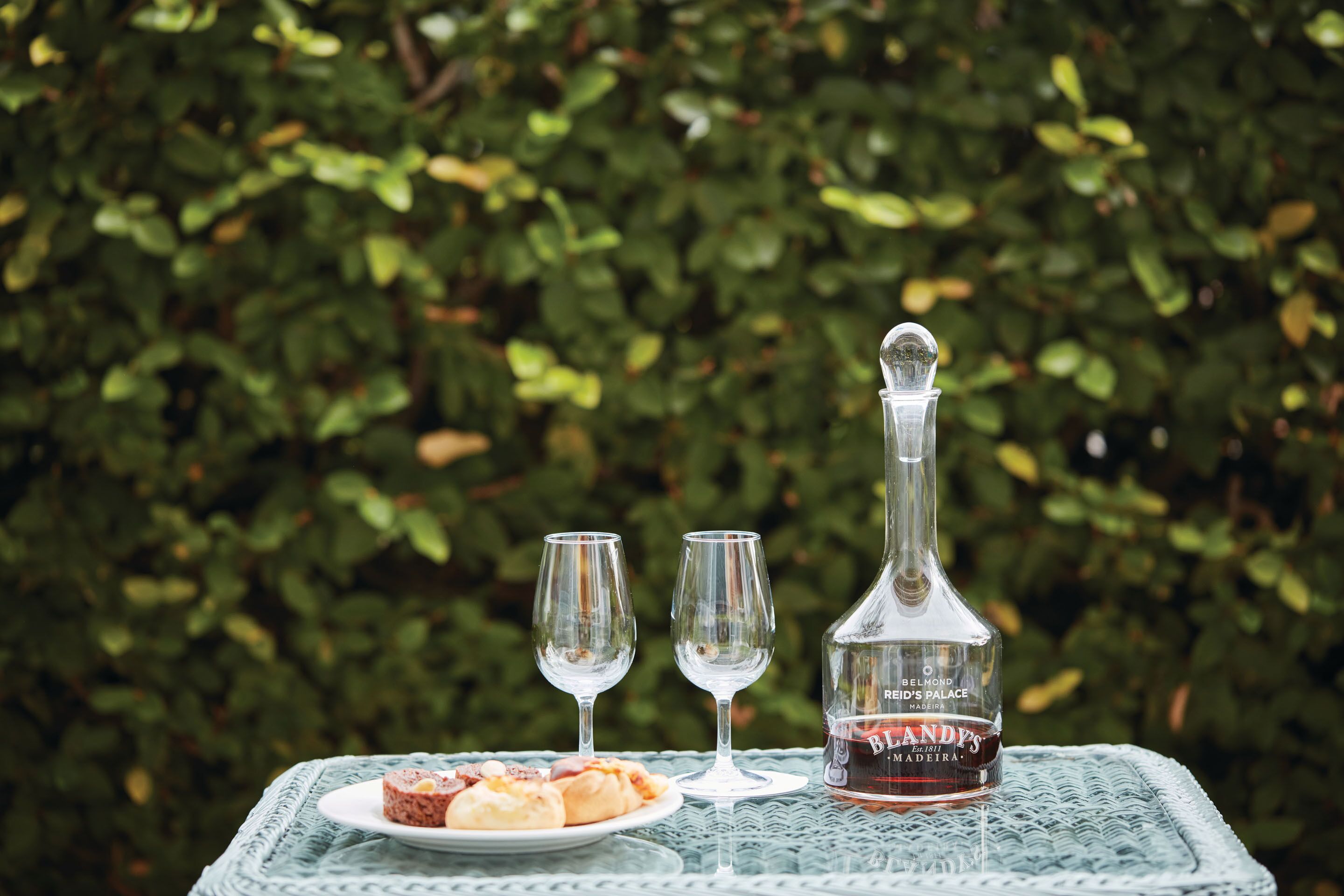 Una botella de Madeira Blandy’s sobre una mesa con dos copas y un plato de comida.