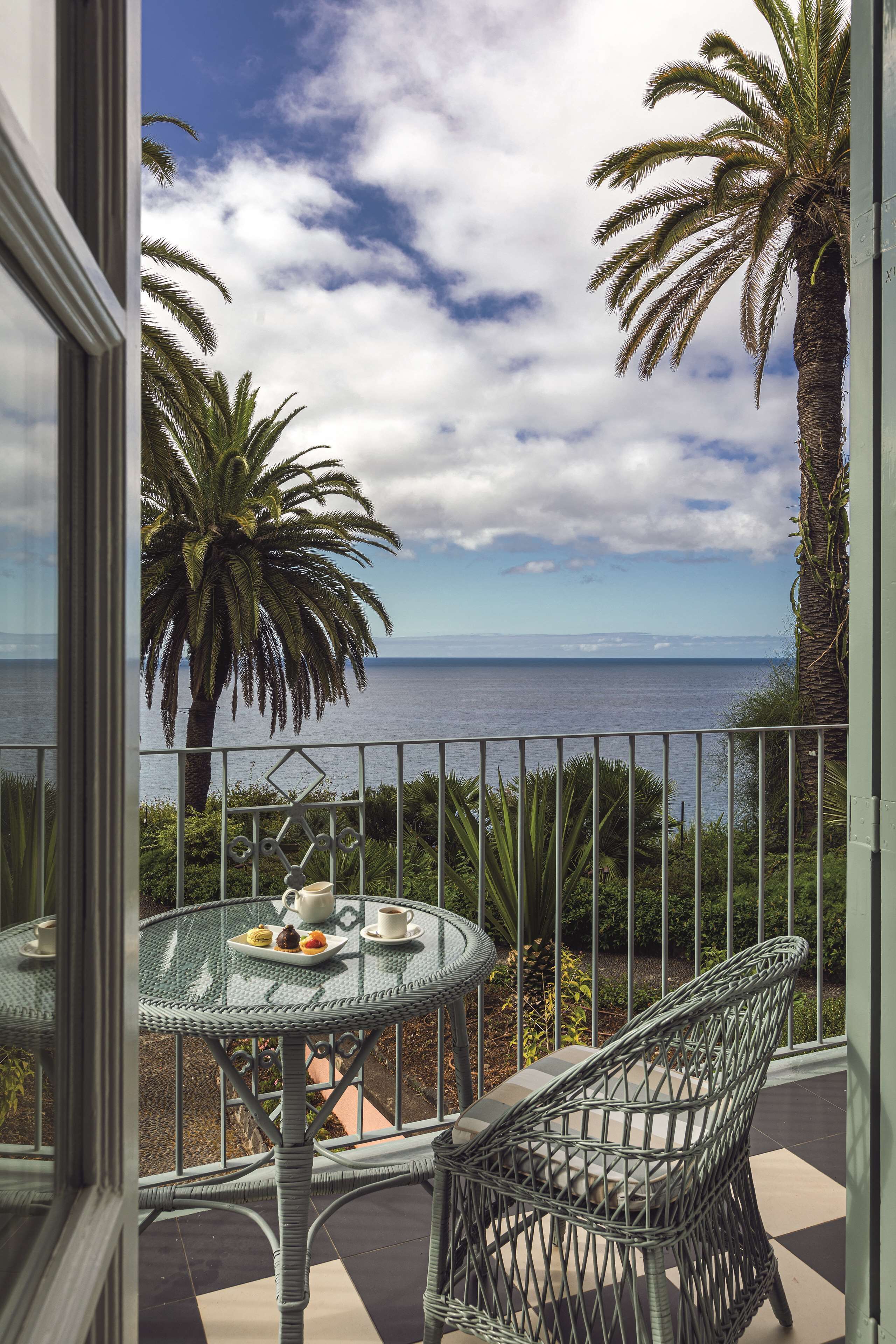 A balcony with a glass table and wicker chairs, overlooking a palm tree and the sea.
