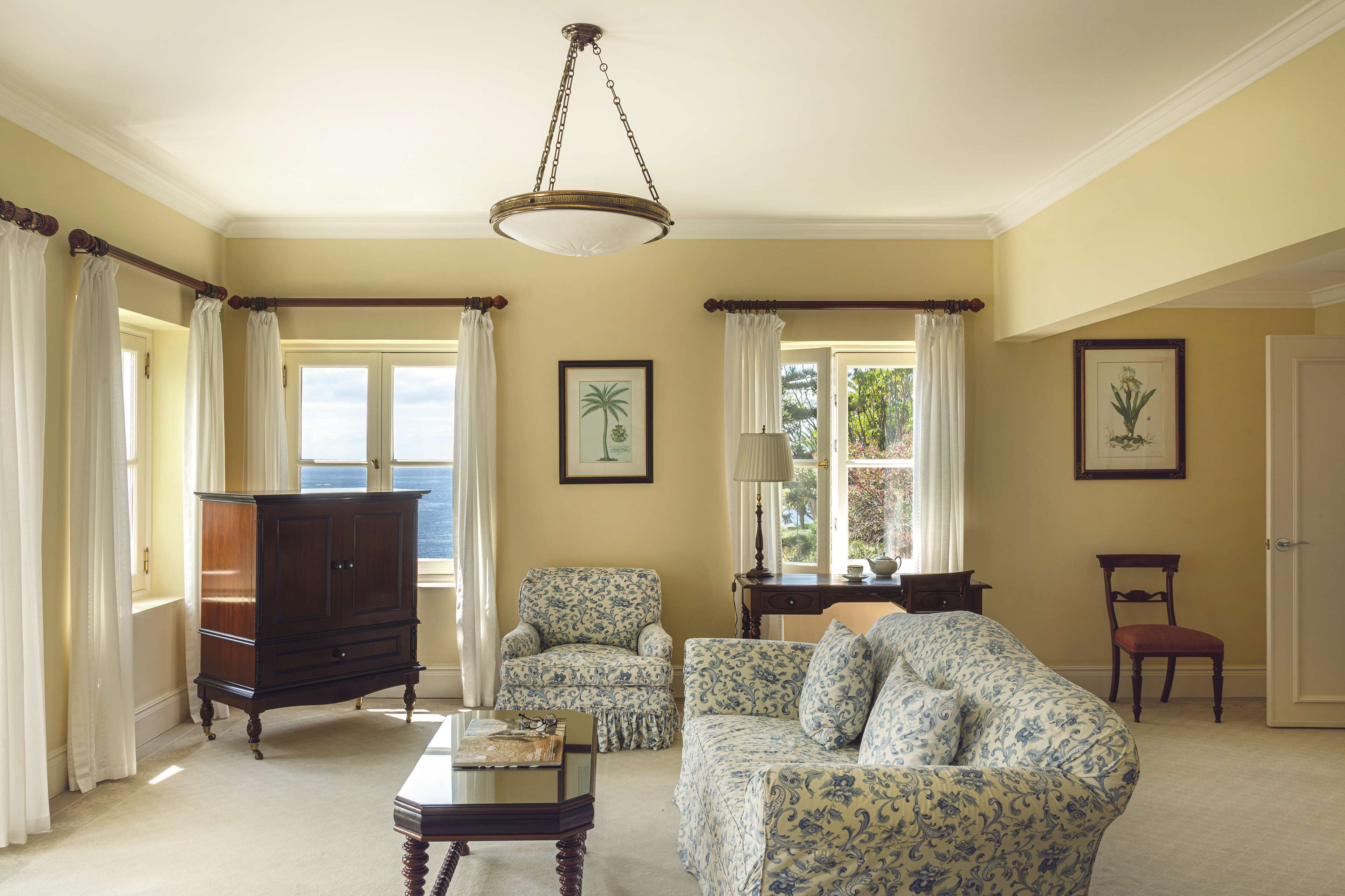 Una habitación con un ventanal grande, una araña de cristal, un armario de madera, un sofá estampado y dos sillones.