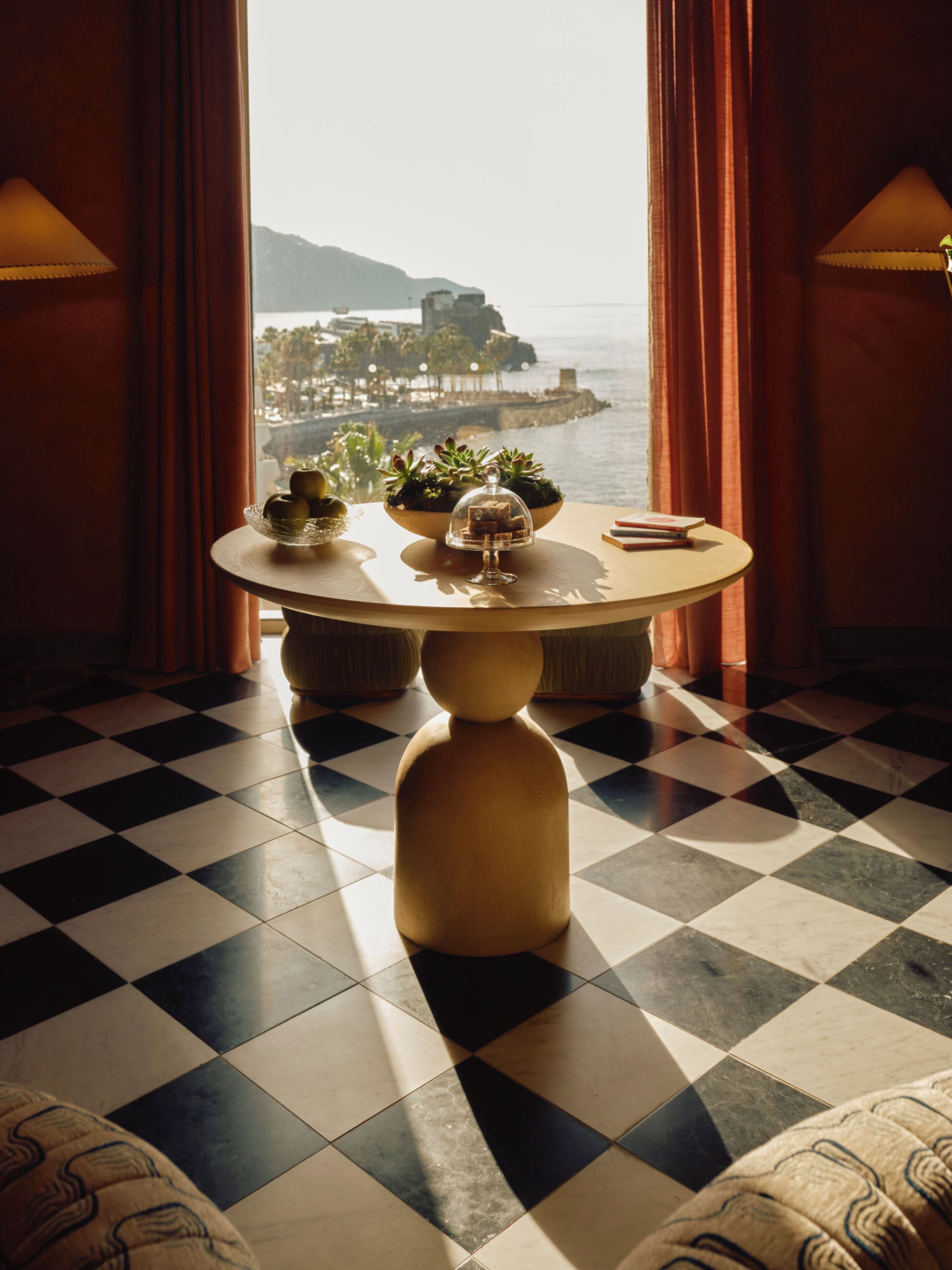 A round table with a checkered floor, a view of a coastal area, and a window with curtains.