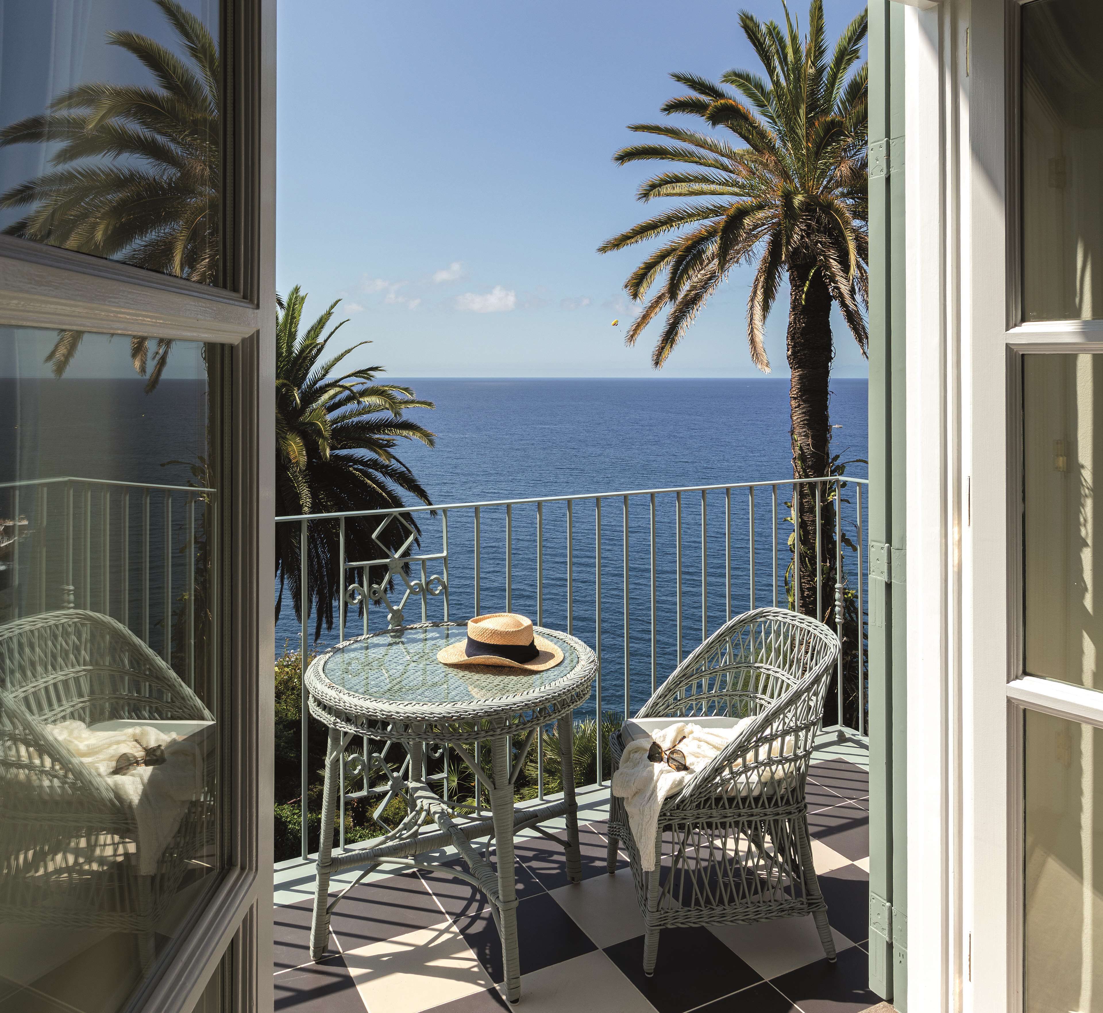 Un balcon avec vue sur l’océan, une chaise en osier, une petite table ronde et un chapeau.