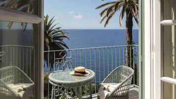 Un balcon avec vue sur l’océan, une chaise en osier, une petite table ronde et un chapeau.