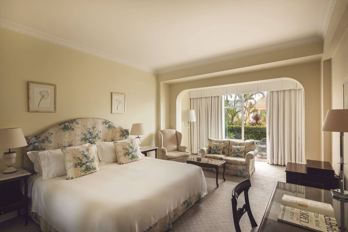 Ruime hotelkamer met een groot bed, een zithoek met een bank en fauteuils, een eettafel en uitzicht op het omliggende landschap door een groot raam.