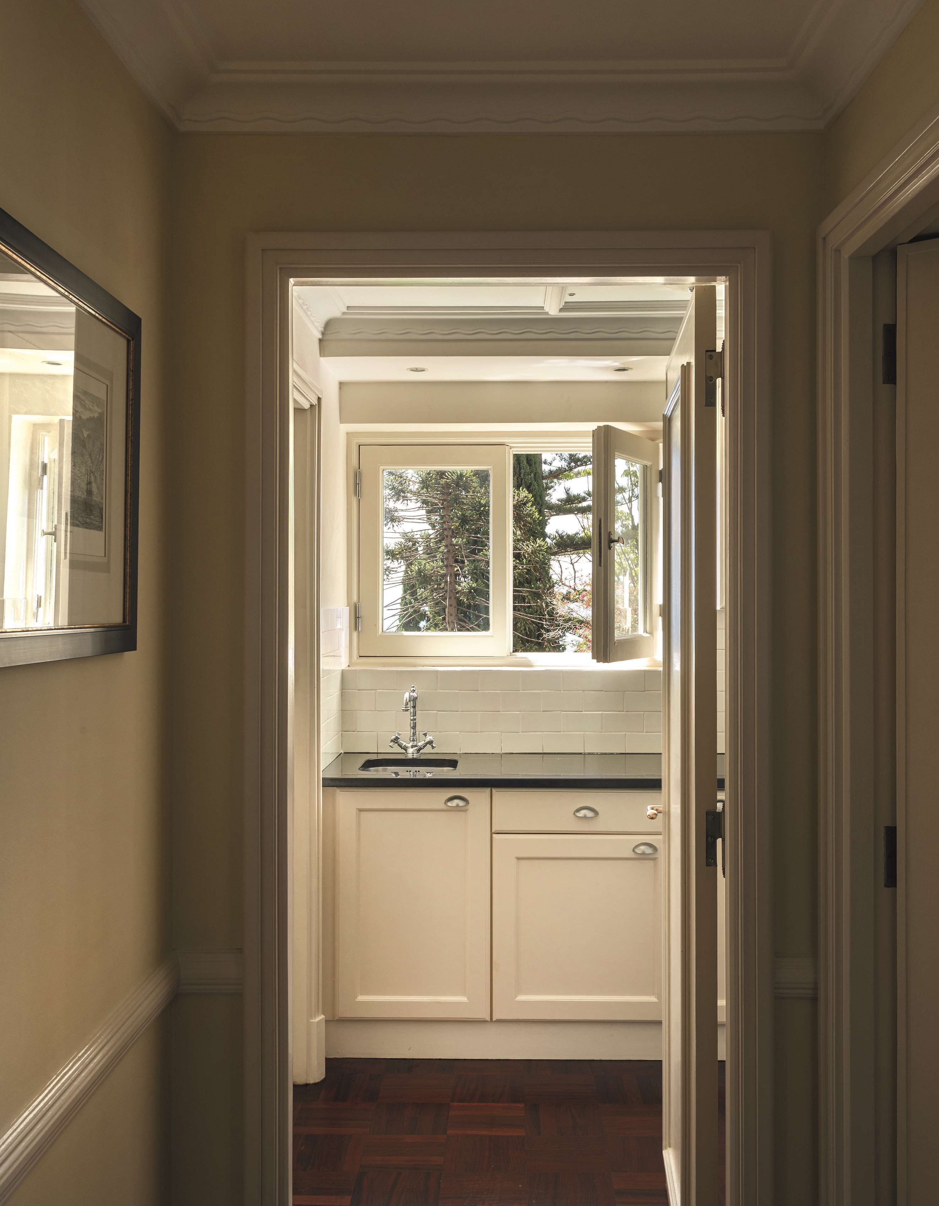 A narrow hallway leading to a kitchen with a window, white cabinets, and a tiled backsplash.