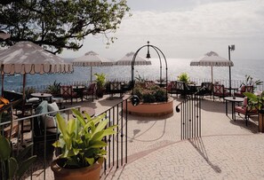 An outdoor seating area with tables and chairs, surrounded by potted plants and umbrellas, overlooking the sea. - Reid's Palace, A Belmond Hotel, Madeira (Funchal)